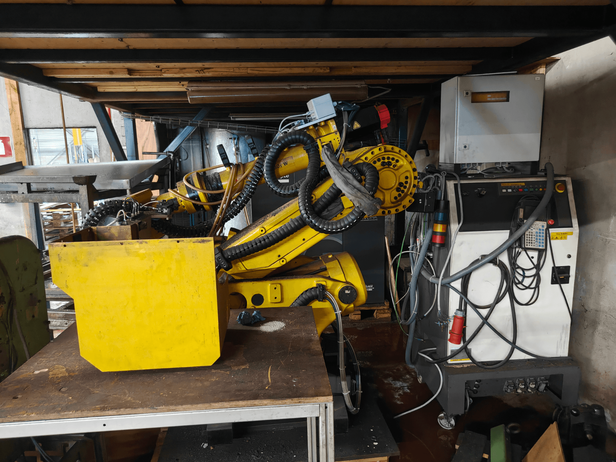 Vista frontal del brazo del robot FANUC S-430i W, de color amarillo, con el panel de control, los cables y un componente amarillo sobre un banco de trabajo.