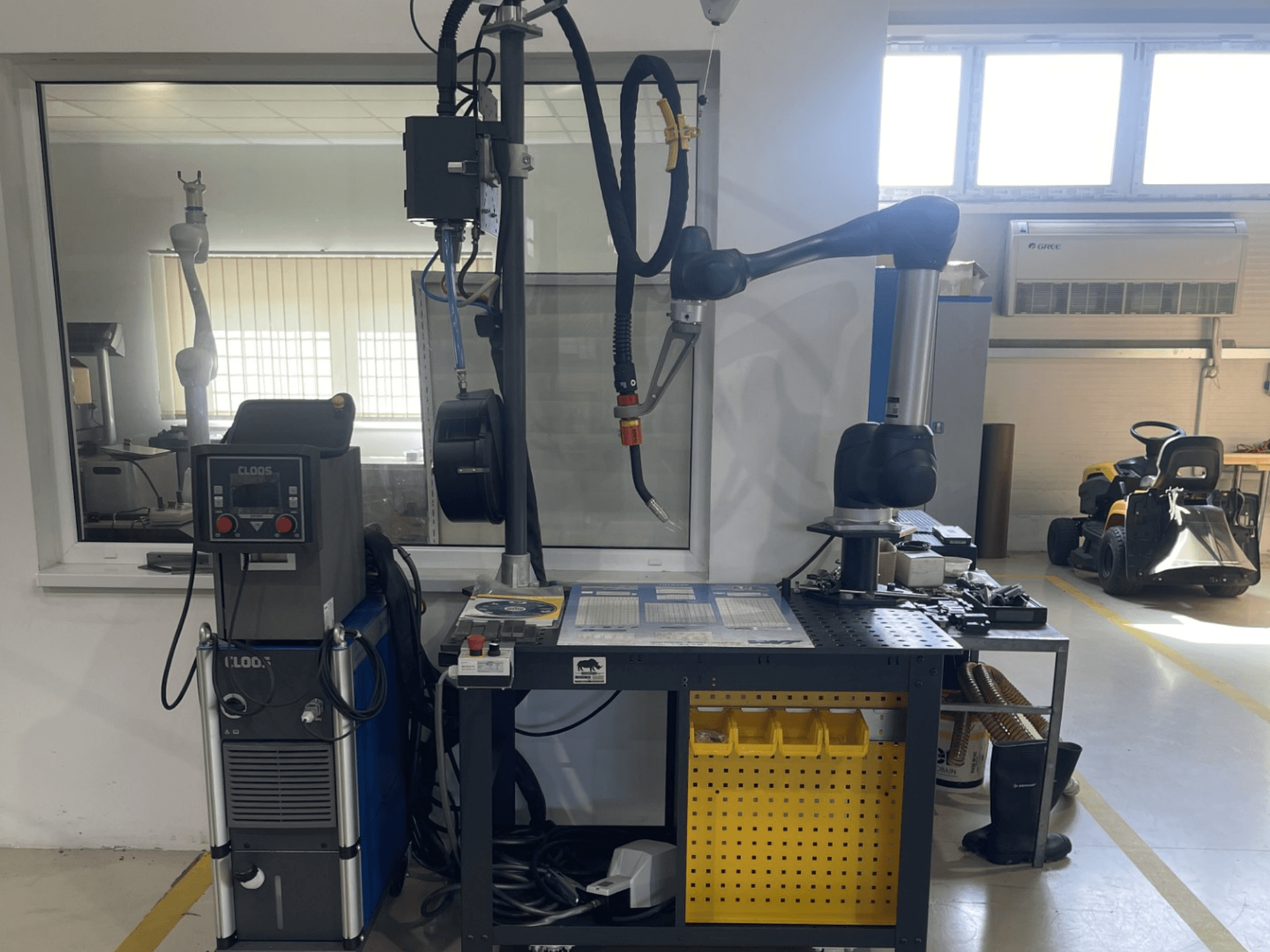 Robot colaborativo DOOSAN M1013 en un taller, vista lateral con equipo de soldadura y herramientas sobre un banco de trabajo.