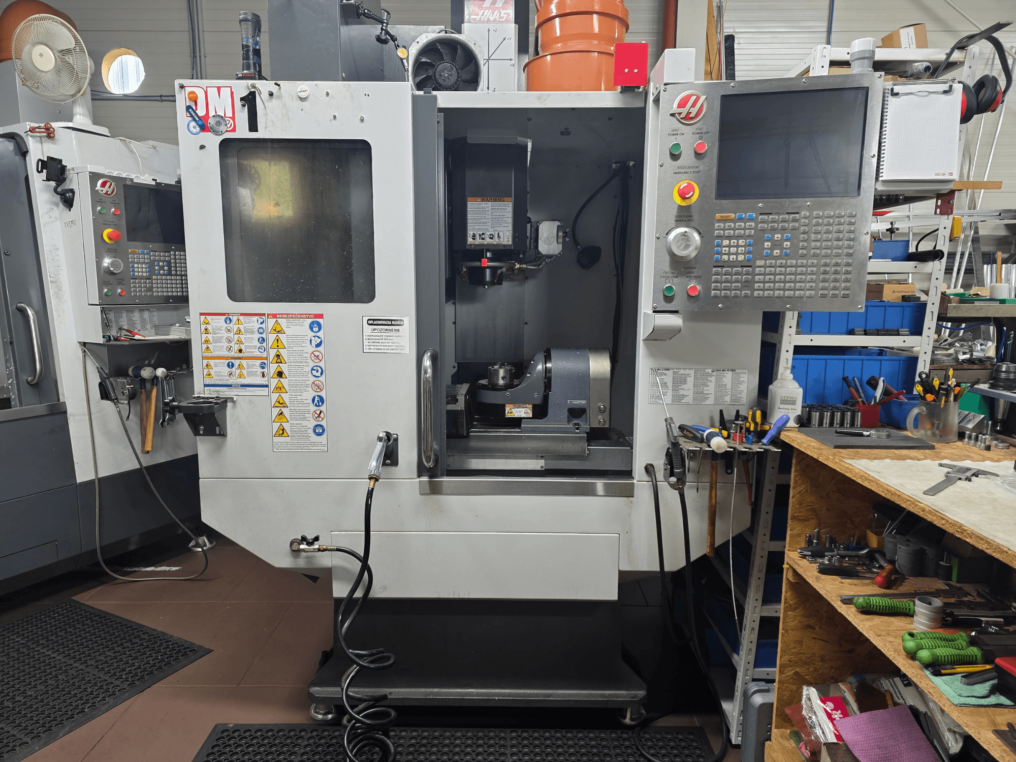 Máquina CNC Haas modelo VF-2, vista frontal, mostrando los controles, la zona del husillo, las etiquetas de seguridad y las herramientas en un banco de trabajo cercano.