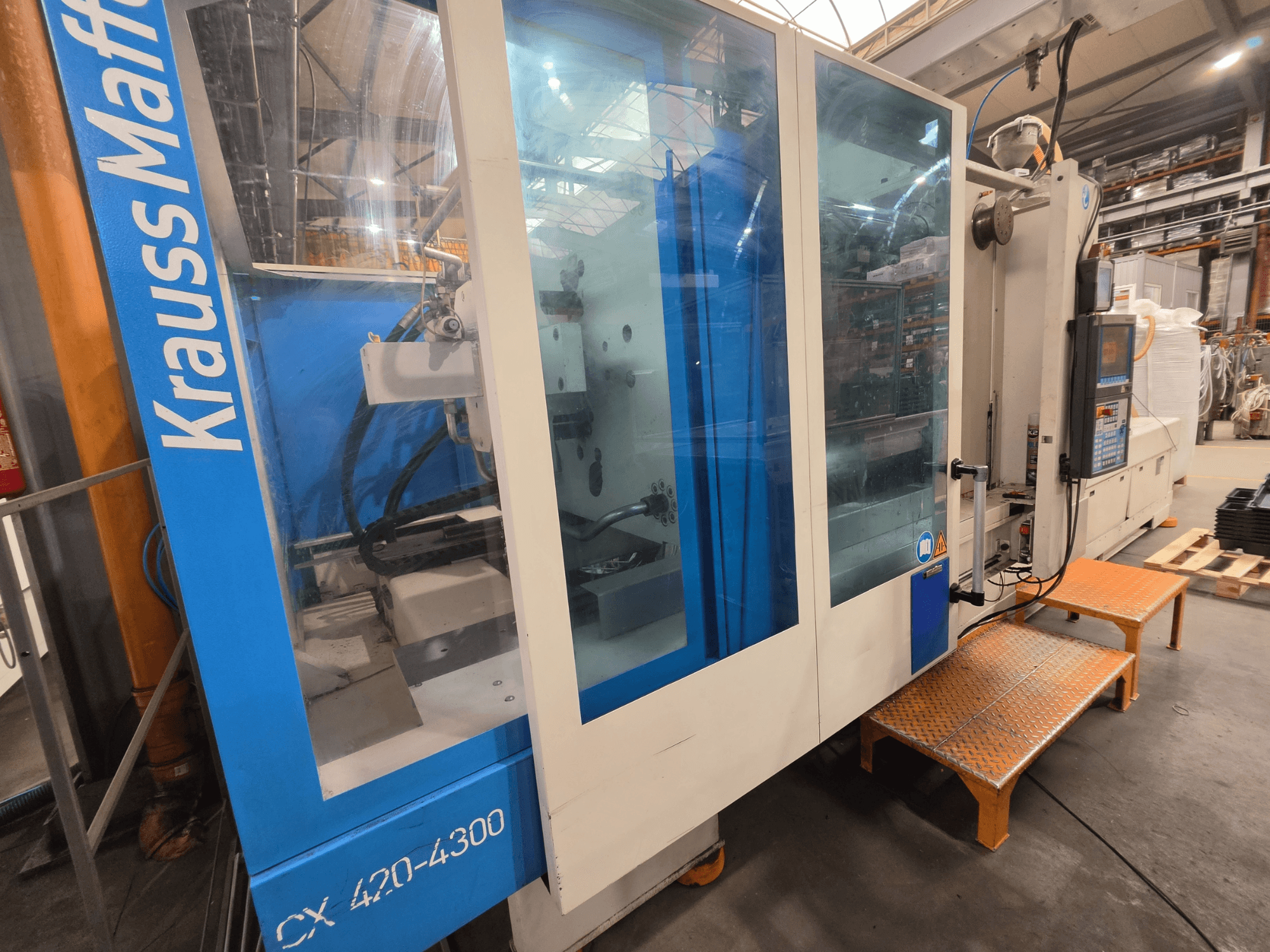 Máquina de moldeo por inyección hidráulica Krauss Maffei CX 420-4300, vista lateral, mostrando el panel de control y los paneles transparentes.