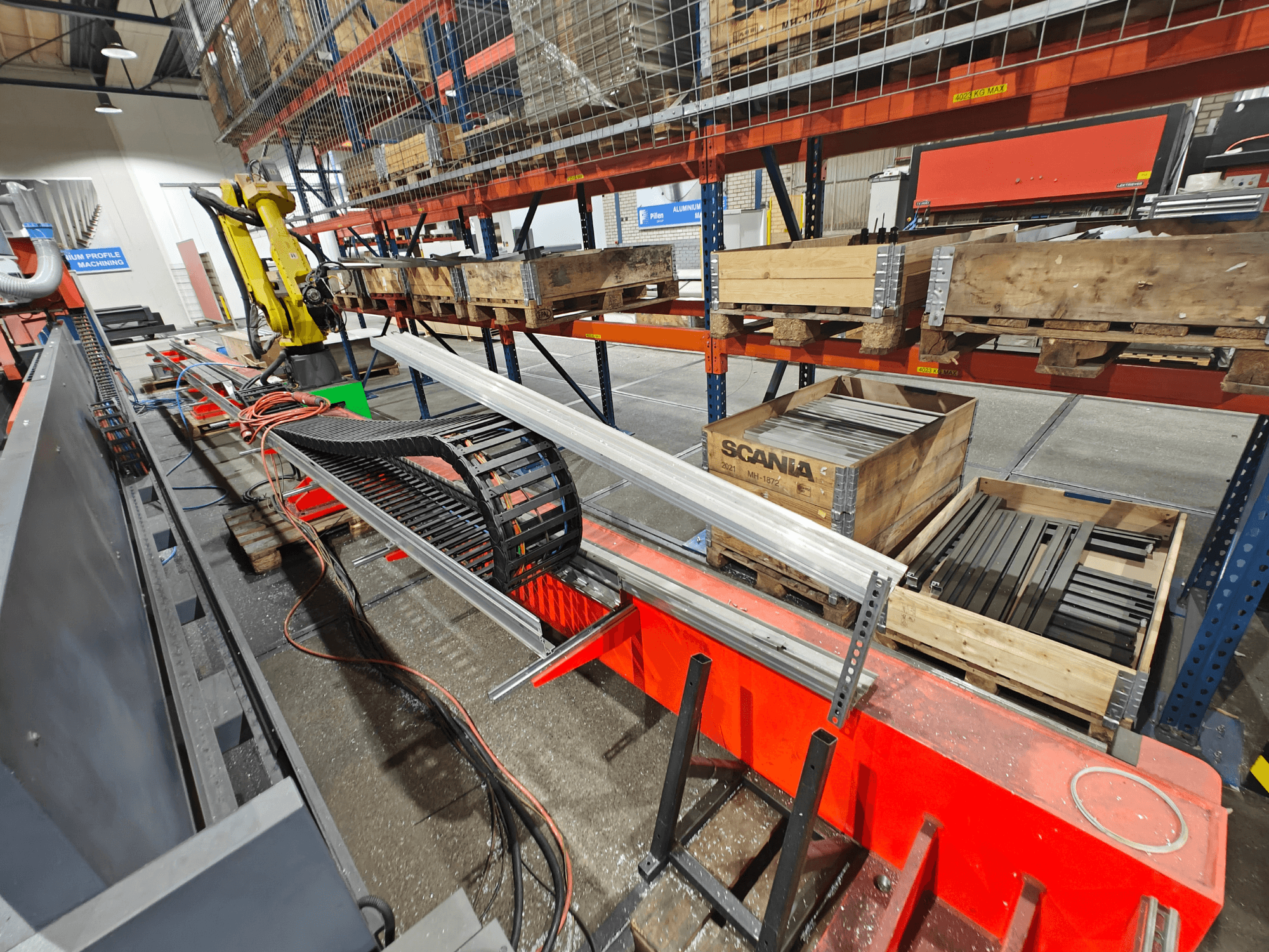 Brazo robótico en estación de trabajo, cables naranja y negro visibles, con cajas Scania y perfiles de aluminio en un entorno de fábrica.