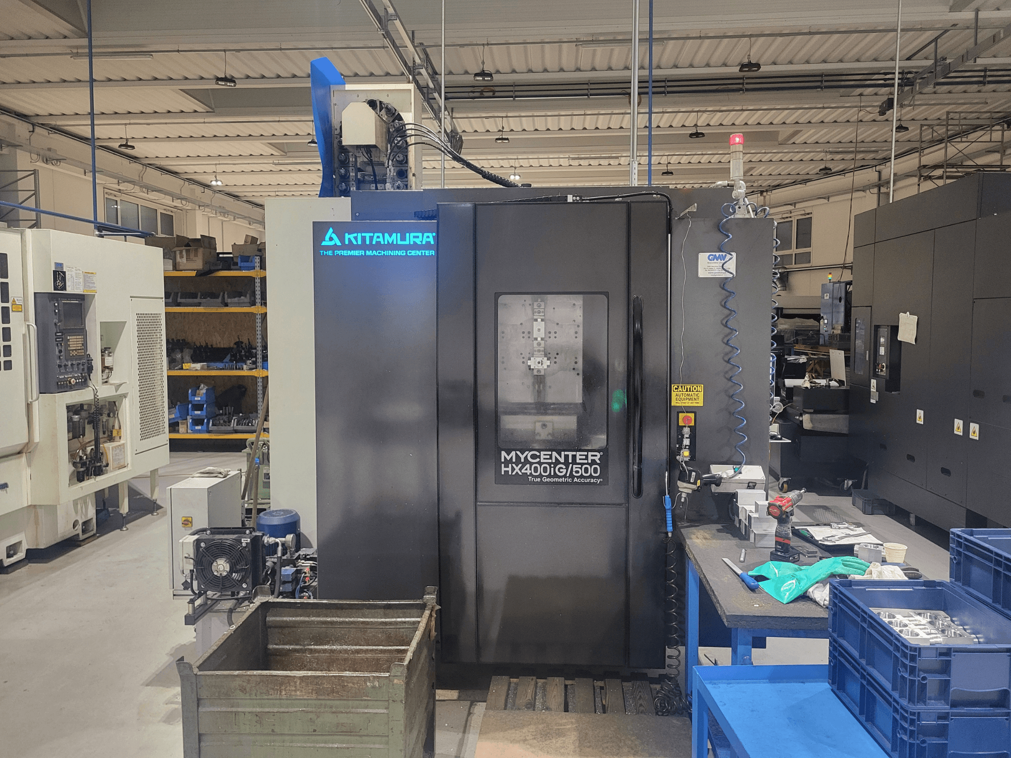Máquina CNC Kitamura Mycenter HX400iG/500 en un taller, vista frontal, con controles, almacenamiento de herramientas y materiales cerca.
