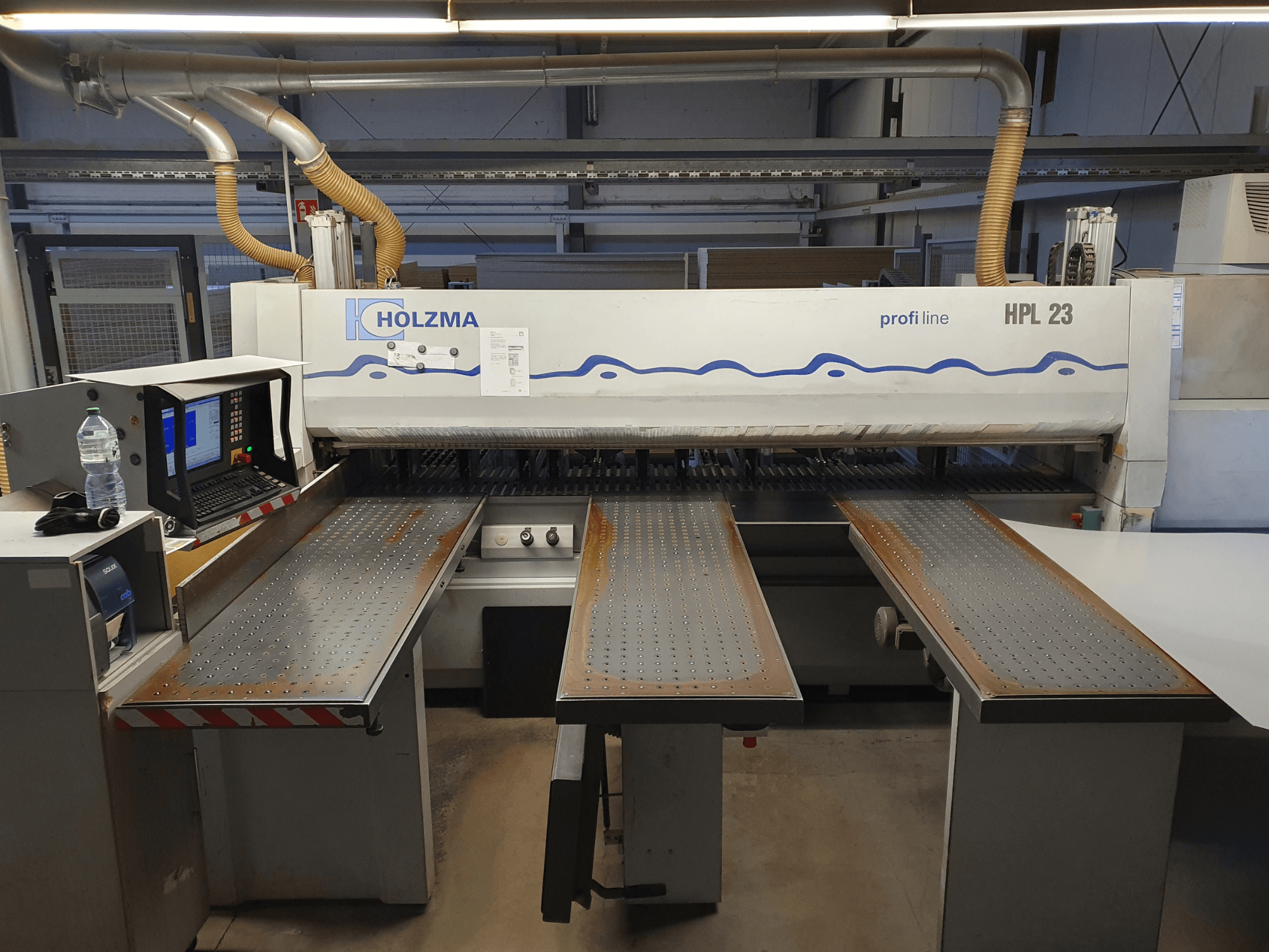 Máquina de corte HOLZMA HPL 23 mostrada desde la vista frontal, con tres mesas perforadas, terminal de ordenador y botella de agua cerca.