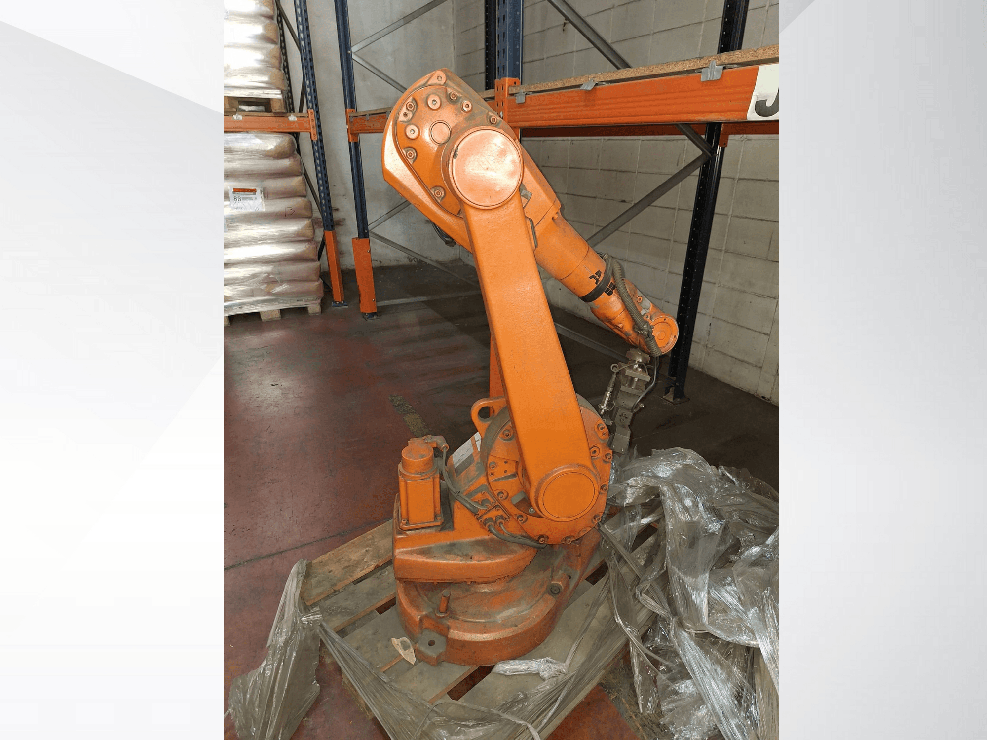 Brazo robótico industrial de color naranja, vista frontal sobre palé de madera, con cable conectado y envoltura de plástico protectora alrededor de la base.