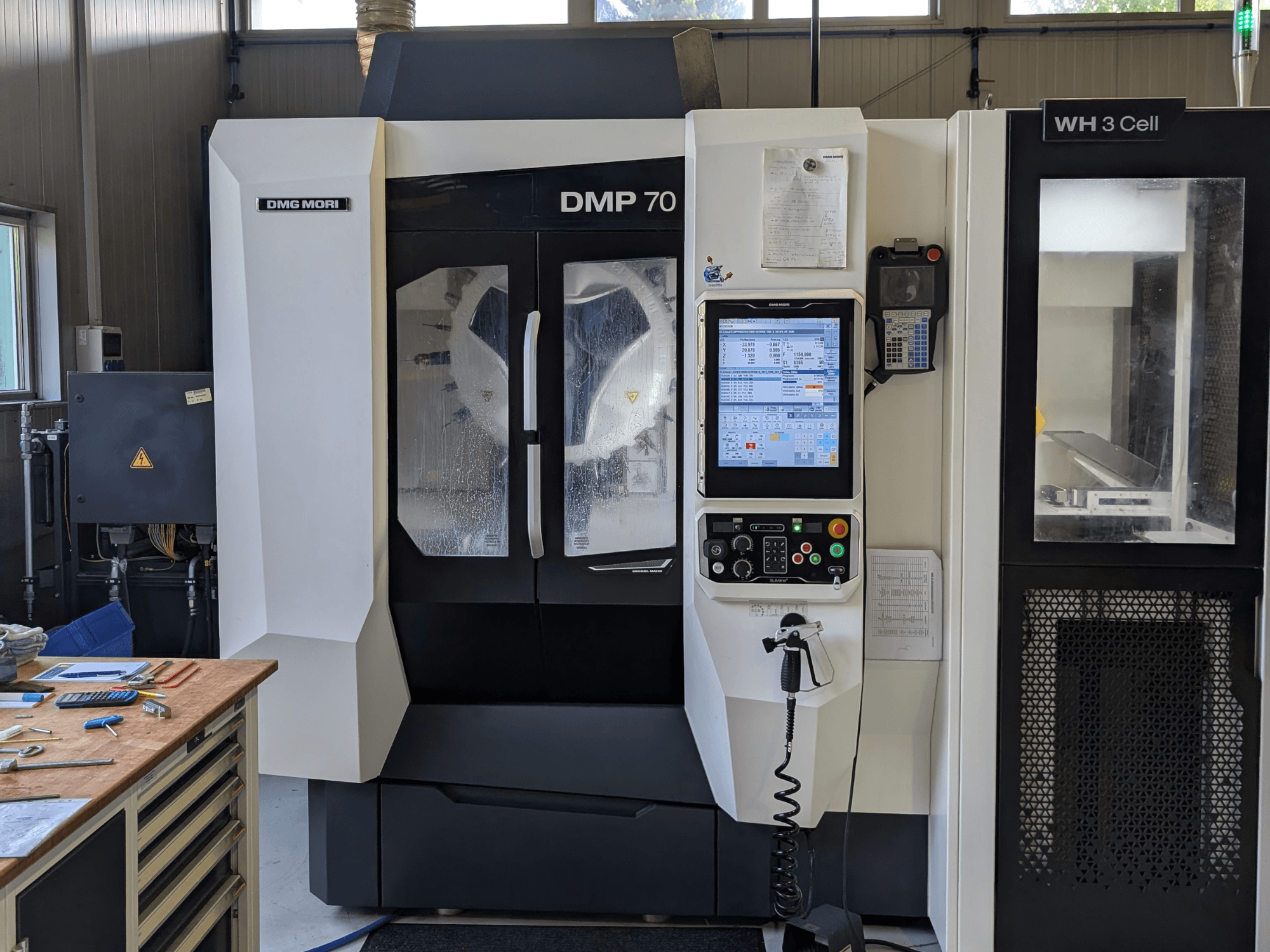 Centro de mecanizado vertical DMG MORI DMP 70, vista frontal con el panel de control y la pantalla de funcionamiento, rodeado de herramientas de taller.