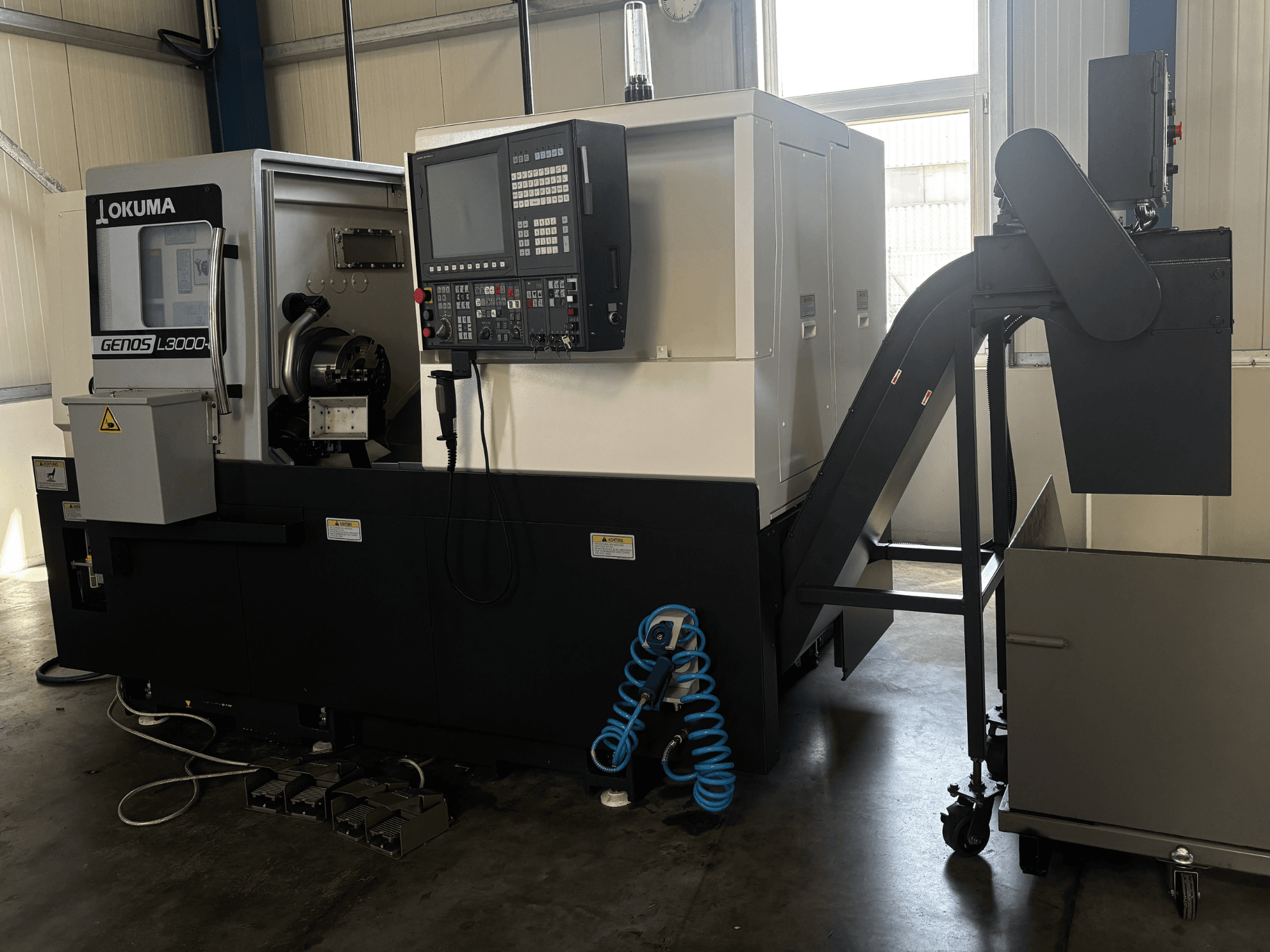Torno horizontal Okuma GENOS L3000-e, vista frontal, en la que se aprecian el panel de control, la cinta transportadora y la zona del husillo.