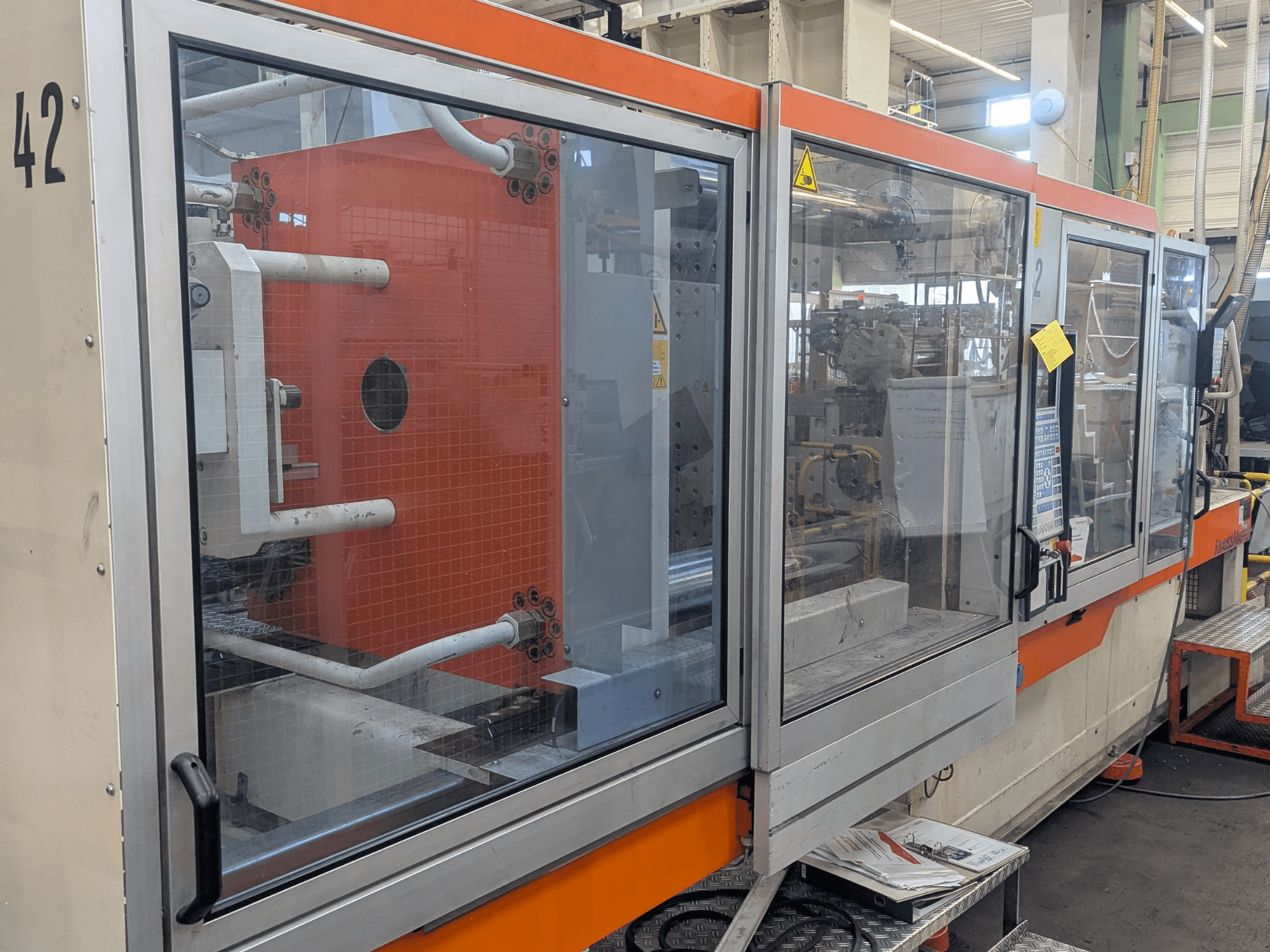 Máquina de moldeo por inyección hidráulica Krauss Maffei KM 350-3000, vista lateral que muestra los controles y los componentes internos detrás del panel de cristal.