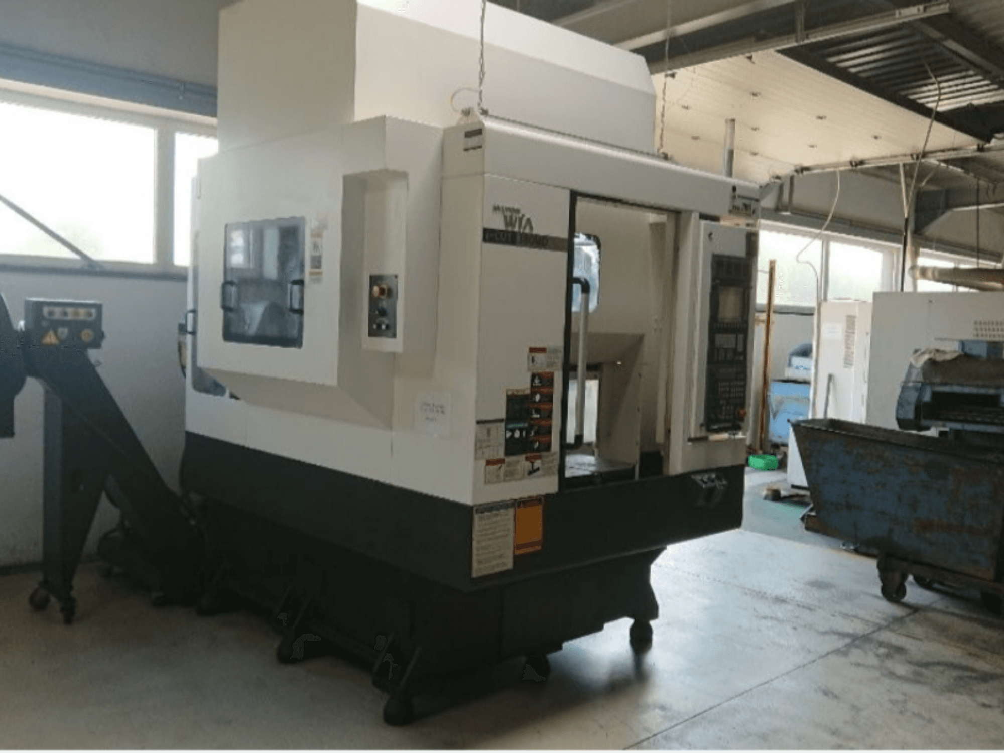 Máquina CNC Haas, vista lateral, con panel de control, etiquetas de advertencia y una rampa de carga, situada en un entorno de taller.