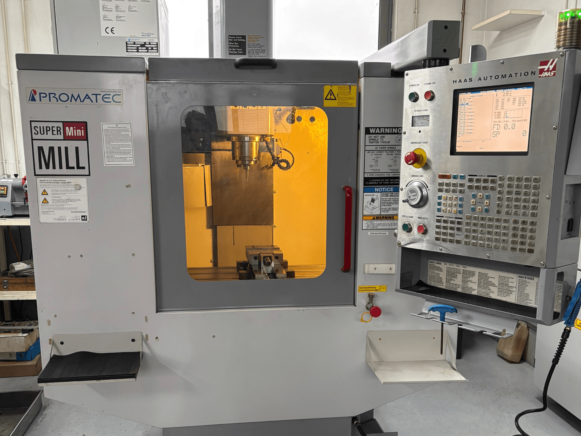 Fresadora Promatec Super Mini en vista frontal, equipada con un panel de control de Haas Automation y herramienta de corte visible en el interior.