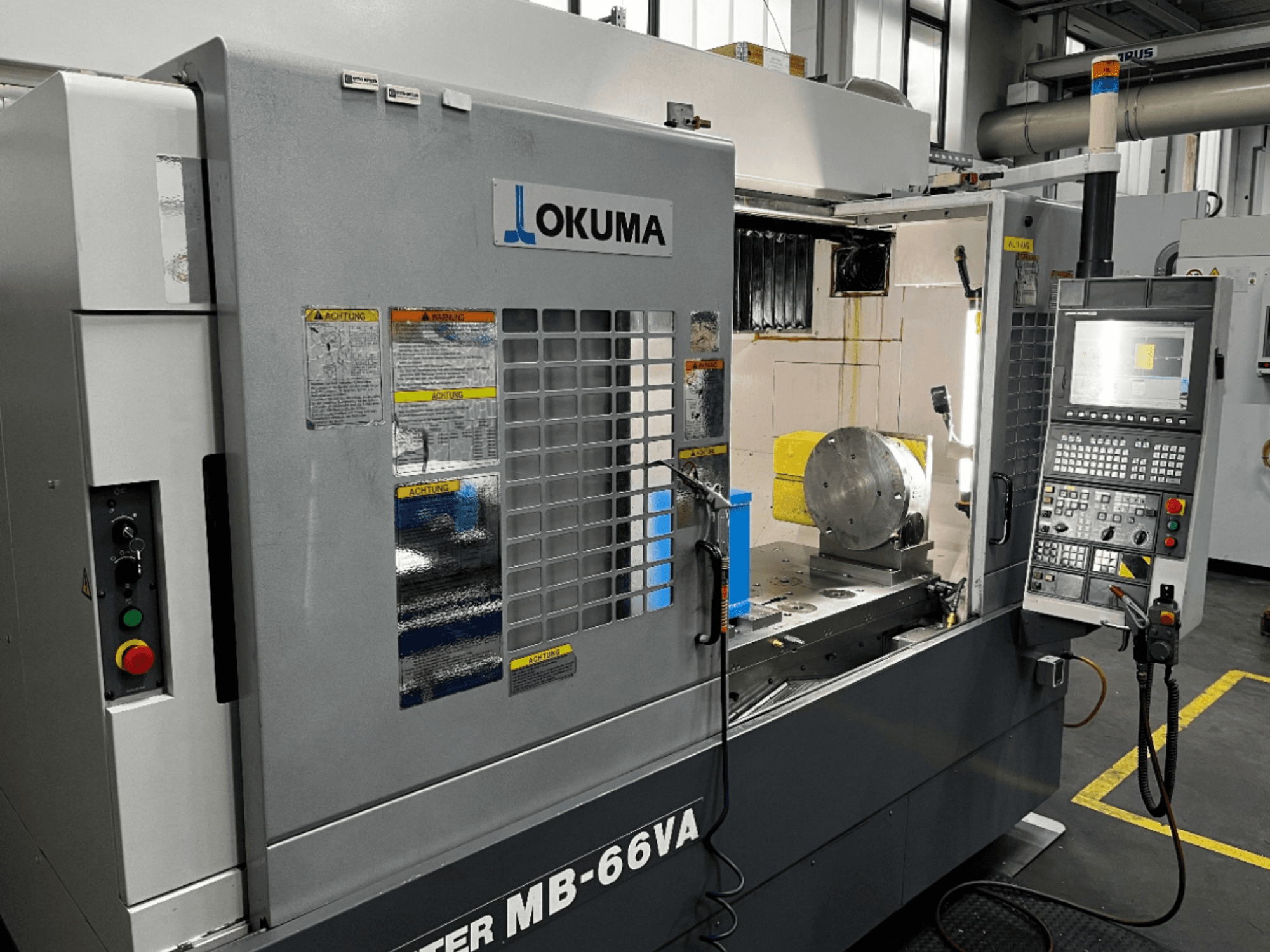 Vista frontal del Centro de Mecanizado Vertical Okuma MB66-VA mostrando el panel de control, la pieza de trabajo y el interior de la máquina con etiquetas de seguridad.