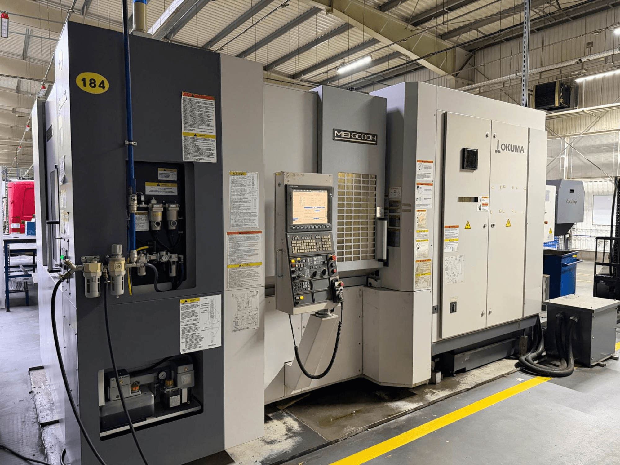 Vista frontal del centro de mecanizado horizontal Okuma MB 5000H, con controles, rotulación y conexiones hidráulicas.