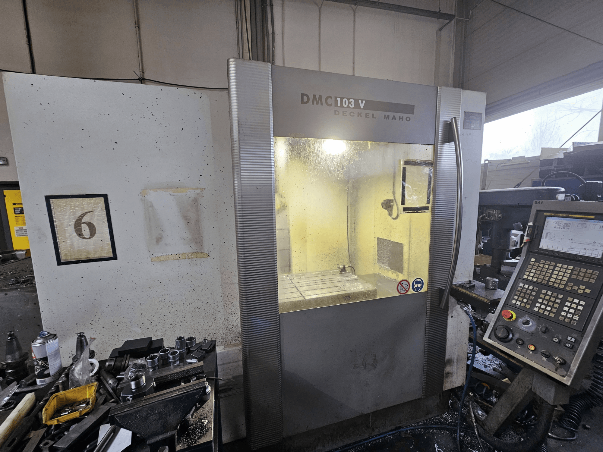 Vista frontal de una máquina CNC Deckel Maho DMC 103 V, mostrando el panel de control, el área de trabajo y el indicador numerado "6".
