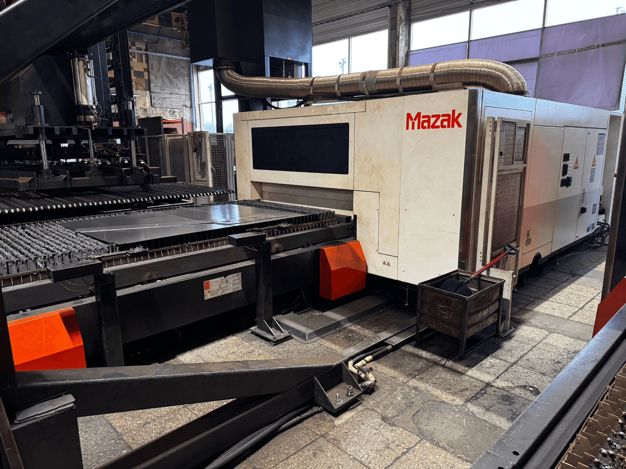 Máquina de corte por láser Mazak en un taller, vista lateral que muestra el panel de control, la cinta transportadora y un recipiente de recogida.