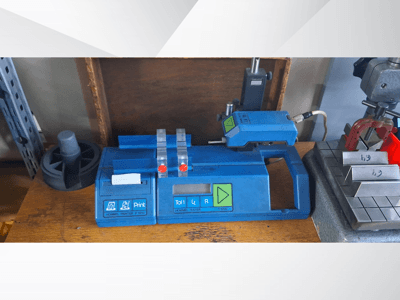 Impresora Hommel T 1000 azul y comprobador T 1 010 con botones rojos, vistos de frente sobre un banco de trabajo con bloques metálicos.