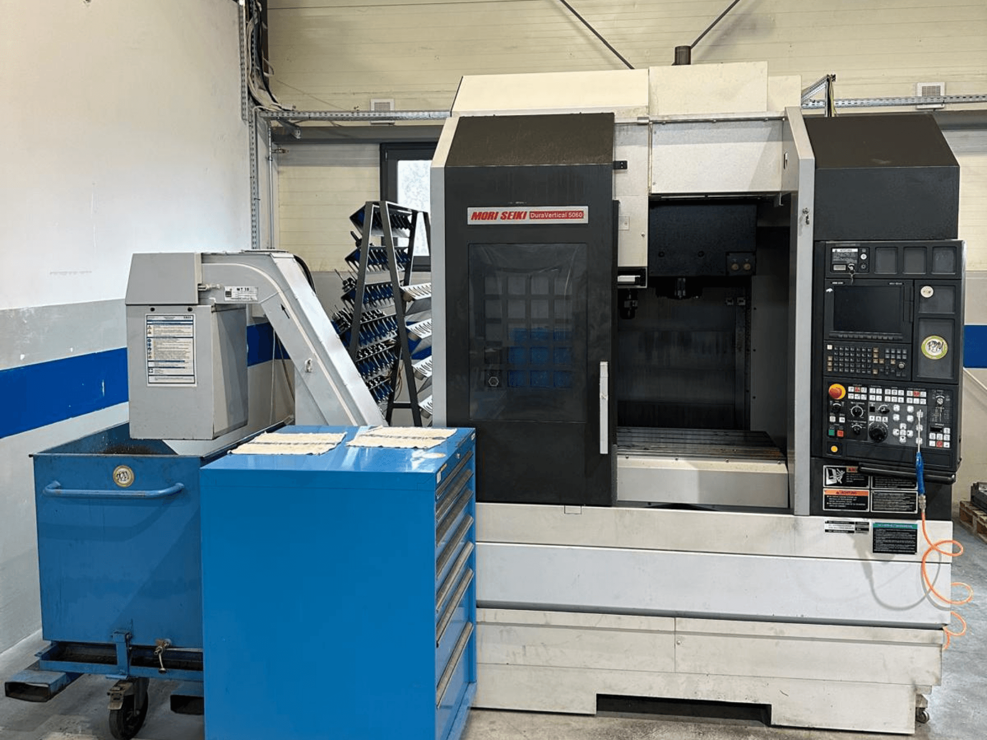 MORI SEIKI DURA VERTICAL 5060, vista frontal de un centro de mecanizado vertical con panel de control y almacén de herramientas adyacente.
