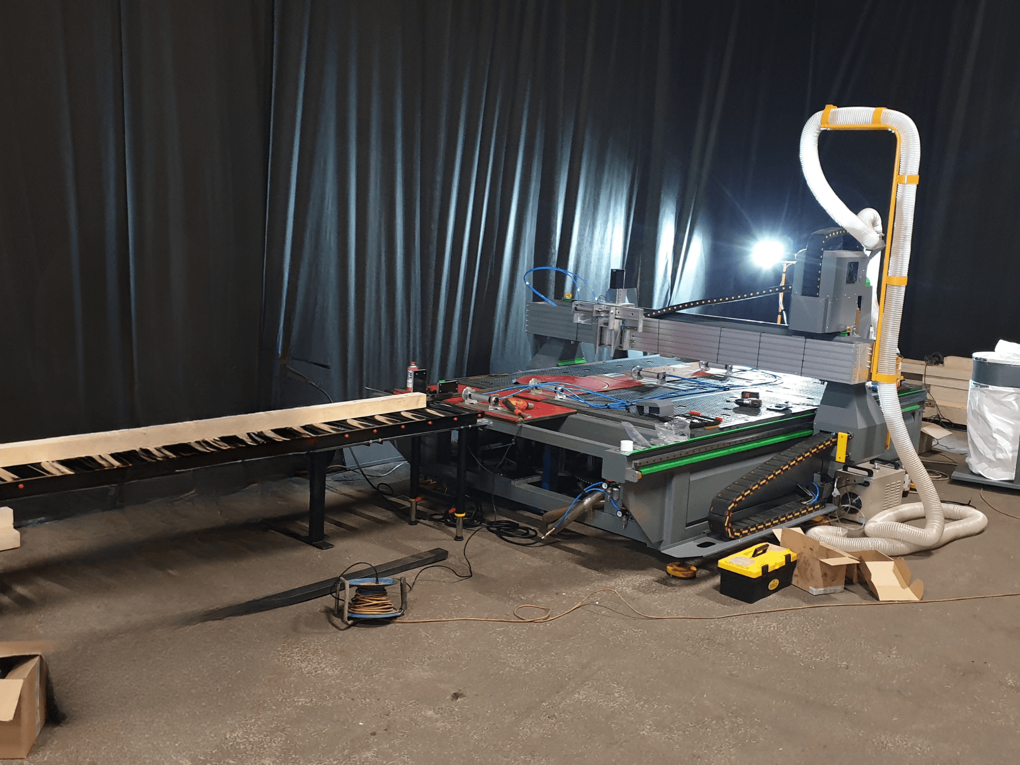 Máquina CNC con transportador lateral, herramientas sobre la mesa y conducto para la recogida de polvo. Fondo negro, vista frontal.