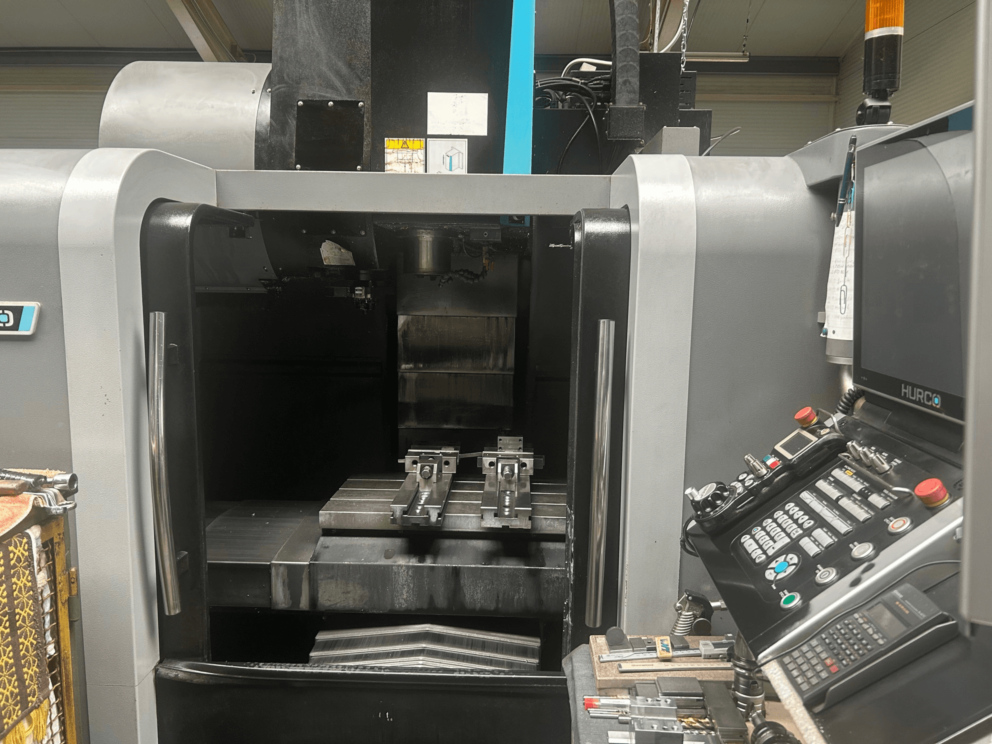 Vista frontal del centro de mecanizado CNC Hurco, mostrando el portaherramientas, la pieza de trabajo metálica y el panel de control con botones y pantalla.