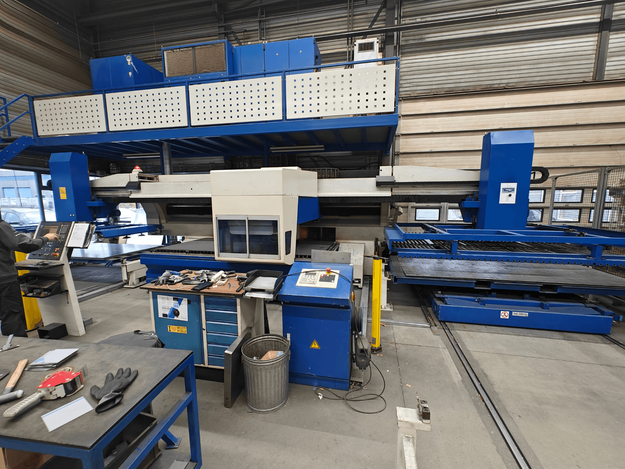 Máquina CNC industrial desde una vista frontal, con construcción en azul y blanco, panel de control y varias herramientas en la mesa de trabajo.