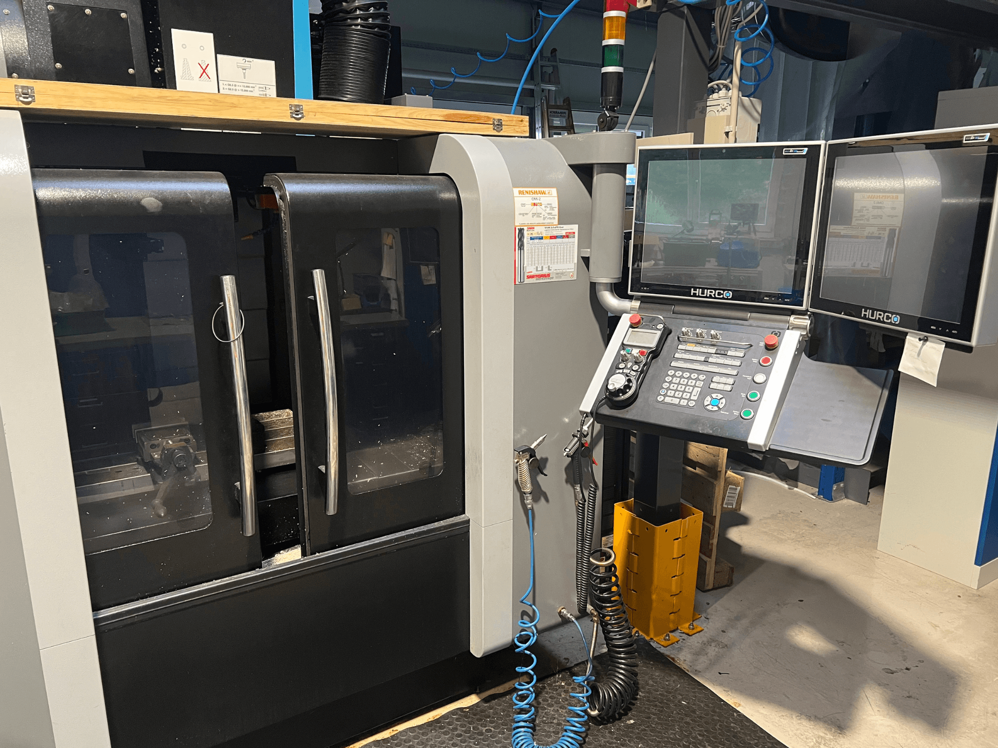 Vista frontal de la máquina CNC Hurco con monitores dobles y panel de control. Puertas transparentes y zona de mecanizado visible en el interior.