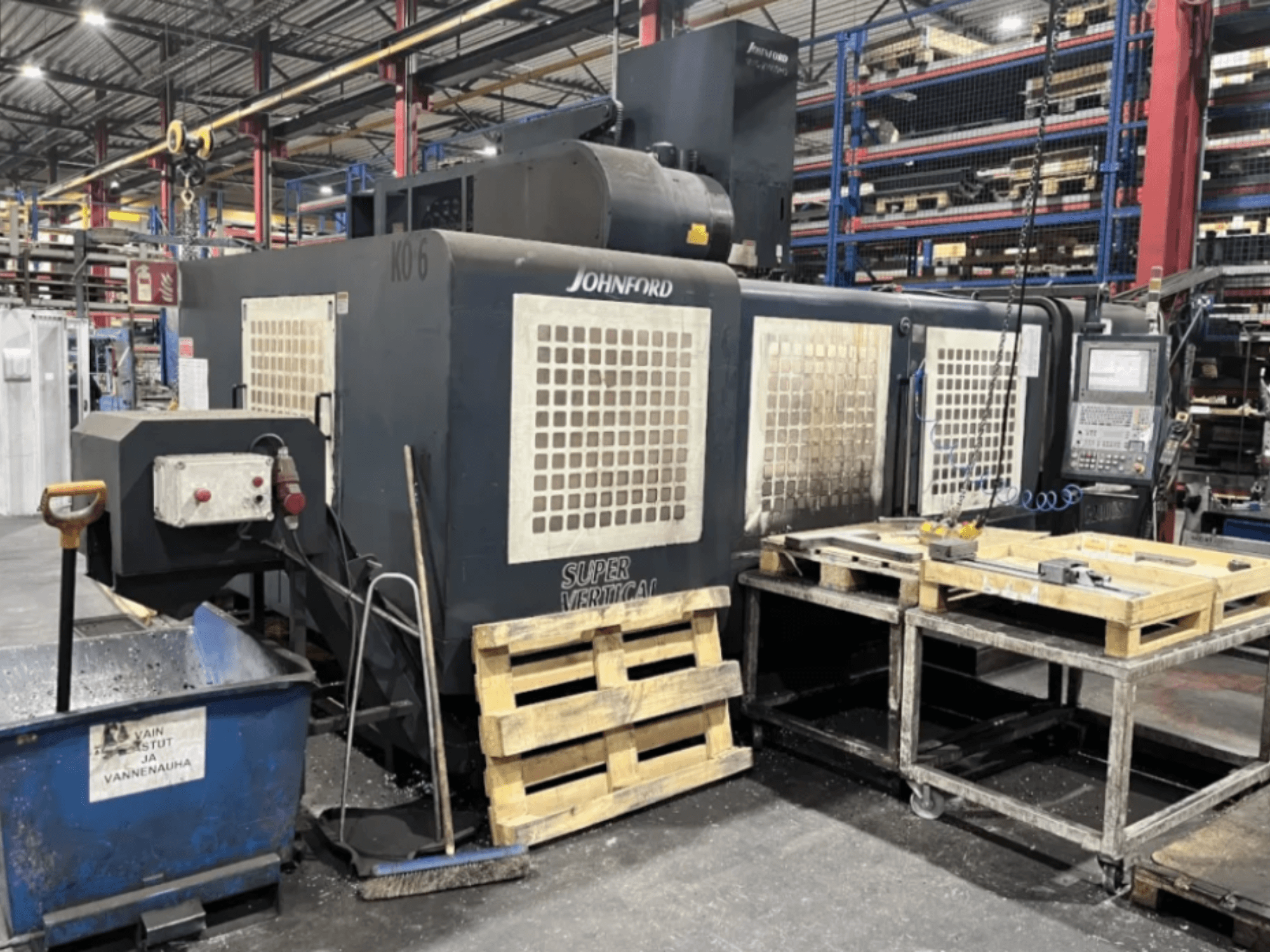 Fresadora Johnford Super Vertical en una fábrica, vista lateral con panel de control, palés y equipo de limpieza cerca.