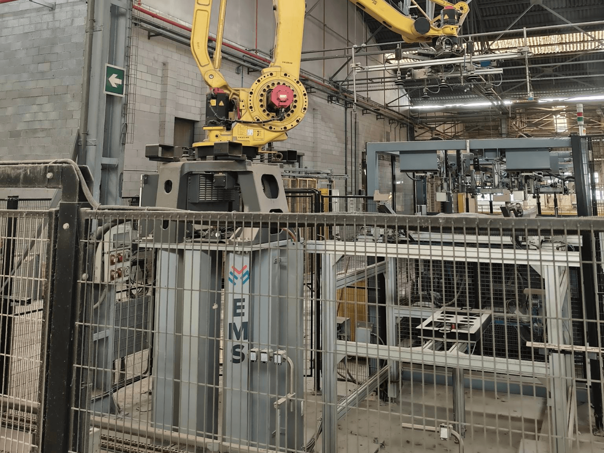 Vista frontal del brazo robótico FANUC M-410iC/315 situado sobre una herramienta EMS, con maquinaria industrial y vallas al fondo.