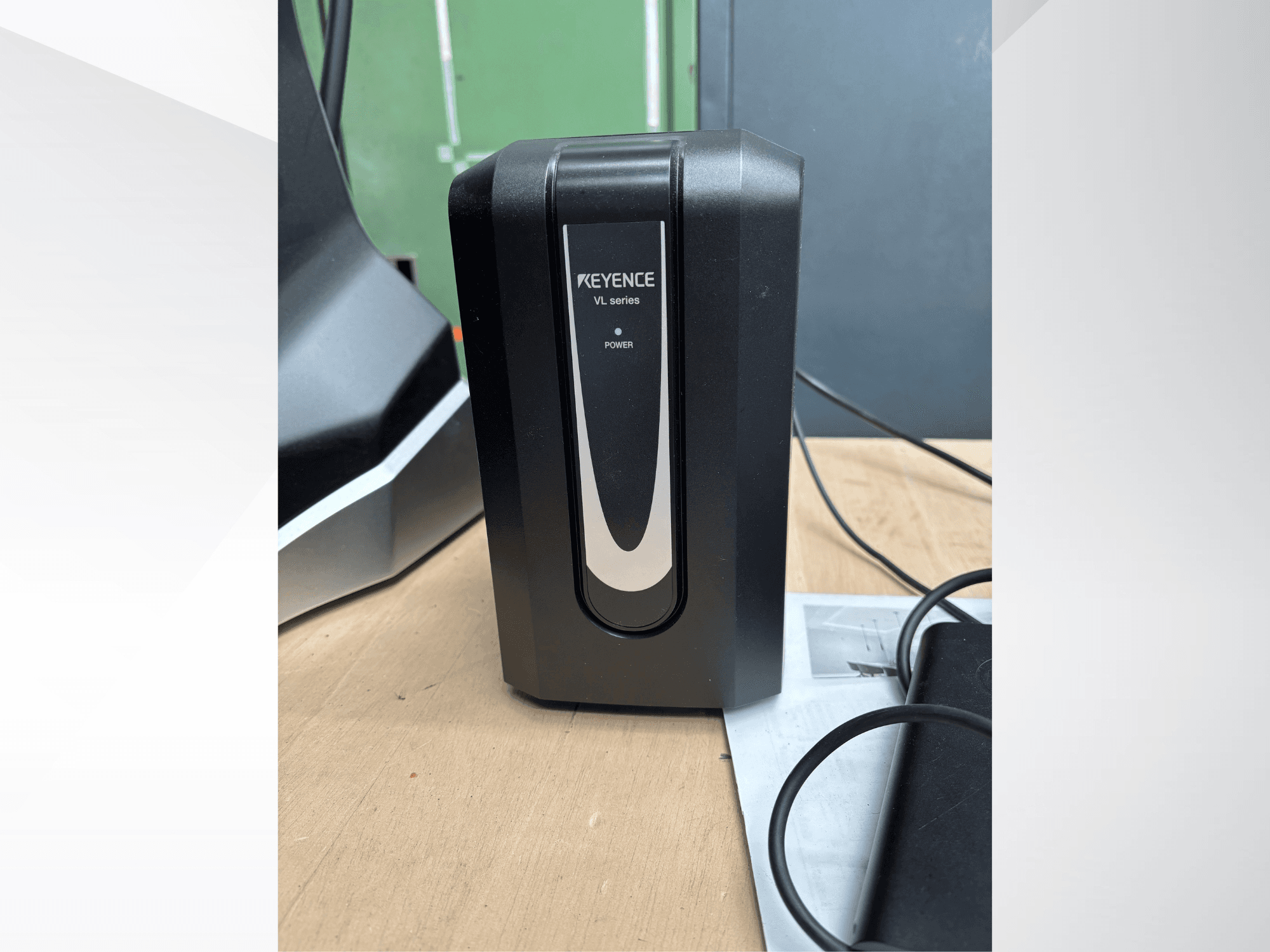 Unidad de alimentación KEYENCE de la serie VL de color negro, vista frontal, con indicador de alimentación y cables conectados sobre una superficie de madera.
