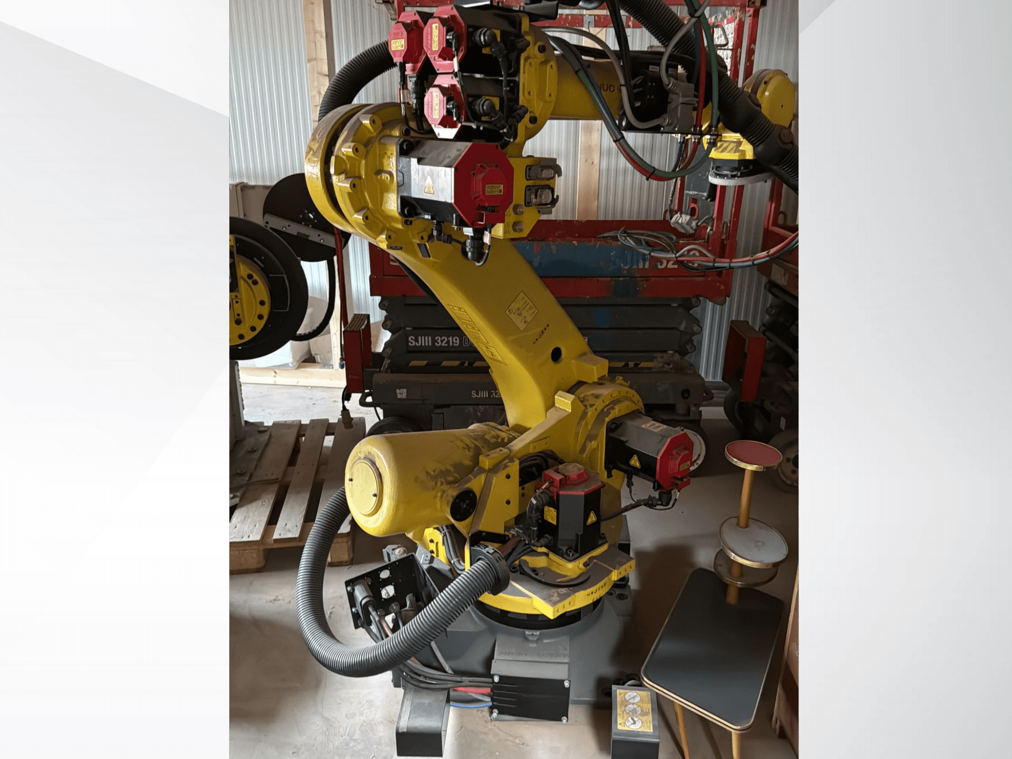 Brazo robótico industrial amarillo con controladores rojos, vista frontal; dispone de múltiples articulaciones y mangueras, colocado en un lugar de trabajo.