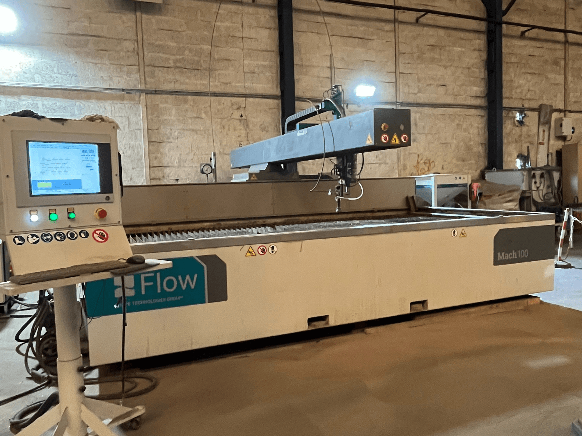 Cortadora por chorro de agua Flow Mach 100, vista lateral, con un panel de control digital y el cabezal de corte visibles sobre una mesa de trabajo.