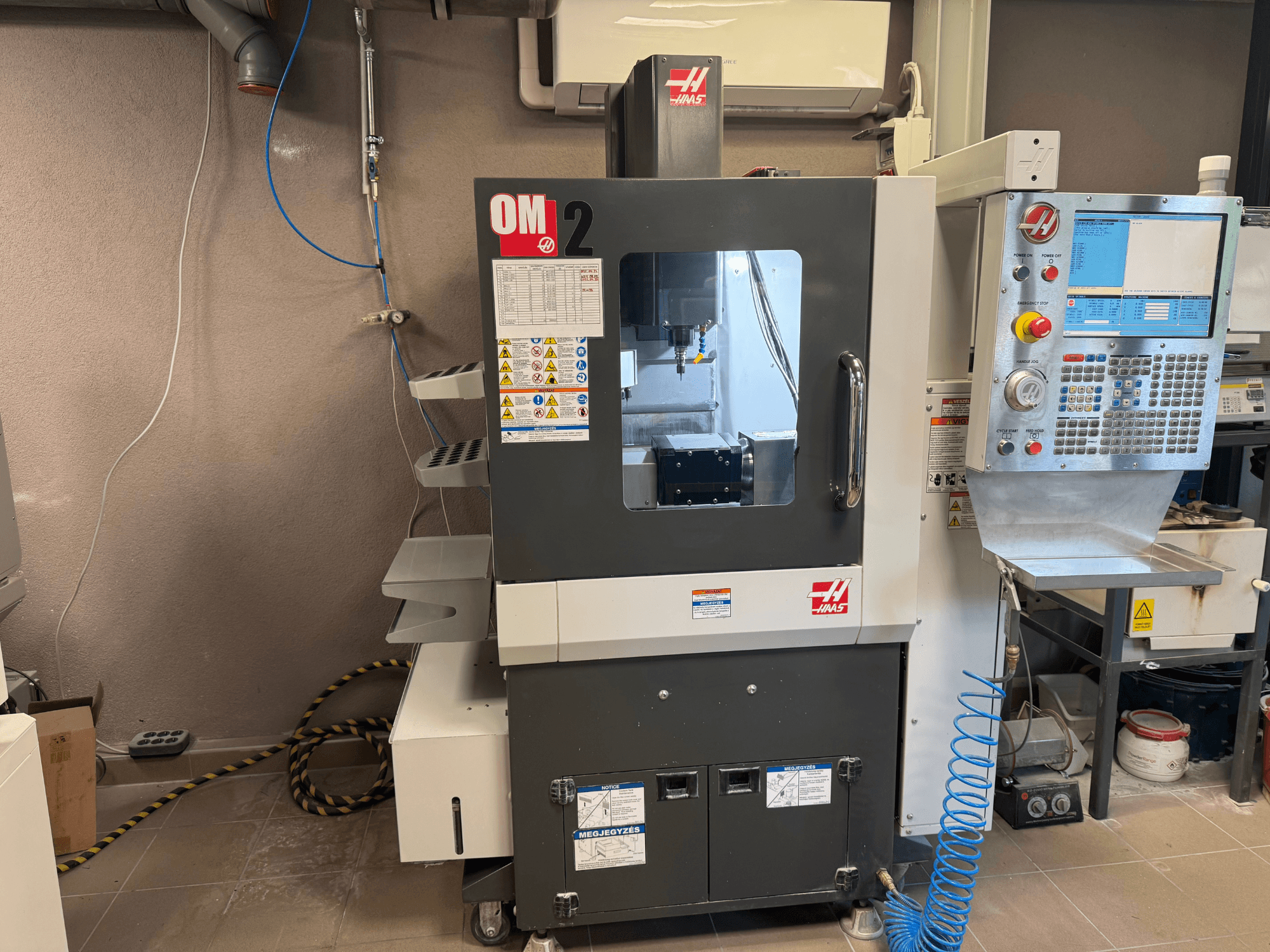 Máquina CNC OM-2 de Haas, vista frontal, con panel de control, husillo y etiquetas de seguridad, colocada en un entorno de taller.