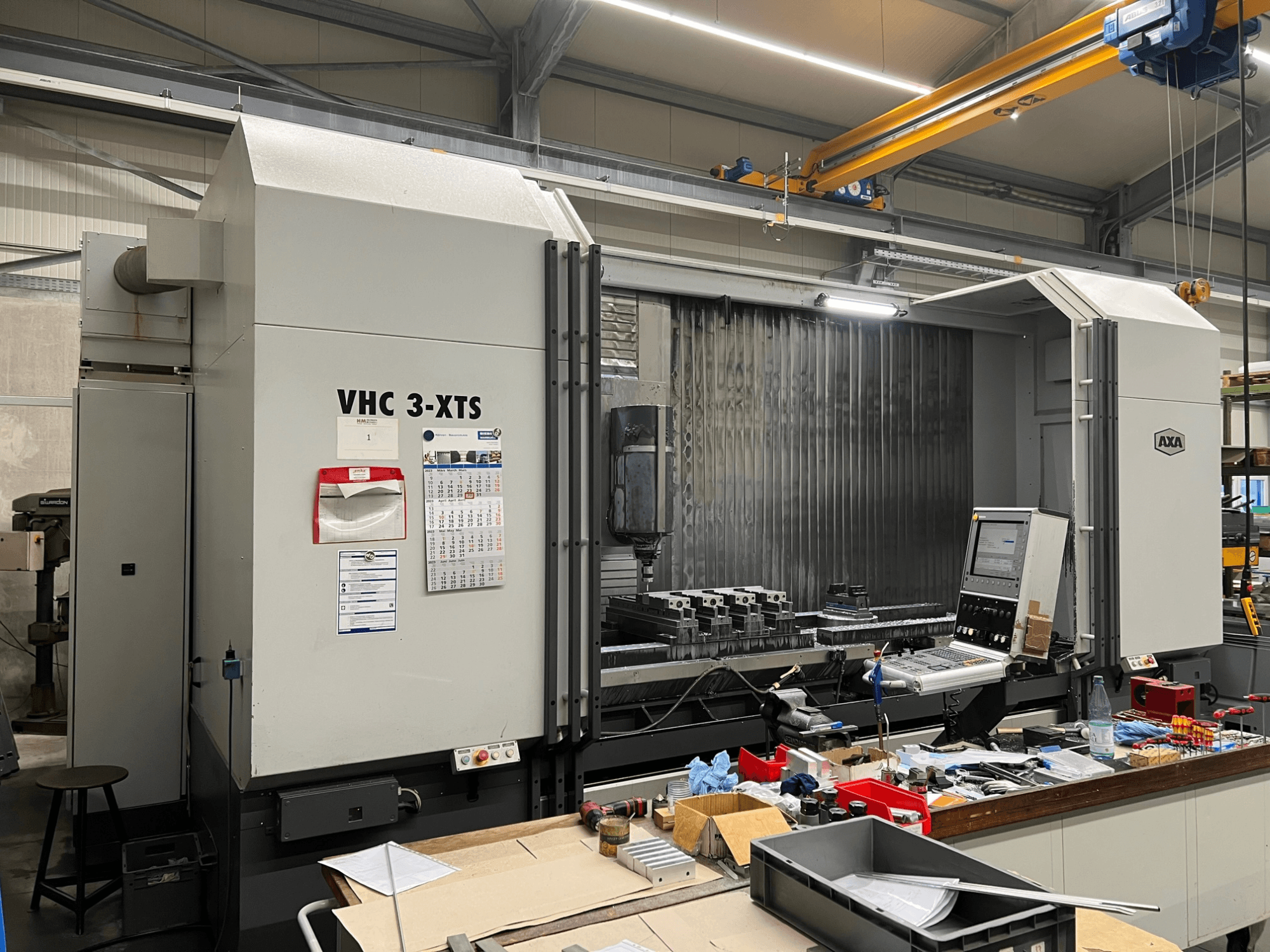 Máquina CNC AXA VHC 3-XTS en vista lateral, con panel de control y área de trabajo abarrotada de herramientas y componentes.