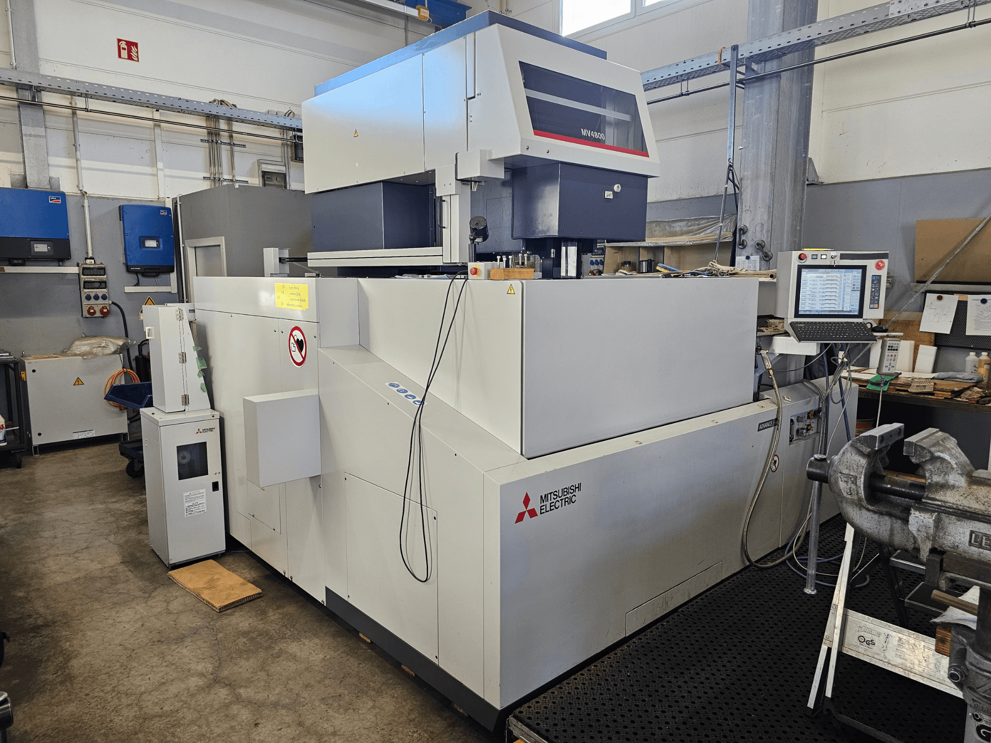 Máquina de electroerosión por hilo Mitsubishi MV4800 en un taller, vista lateral que muestra el panel de control, las etiquetas de seguridad y el equipo circundante.