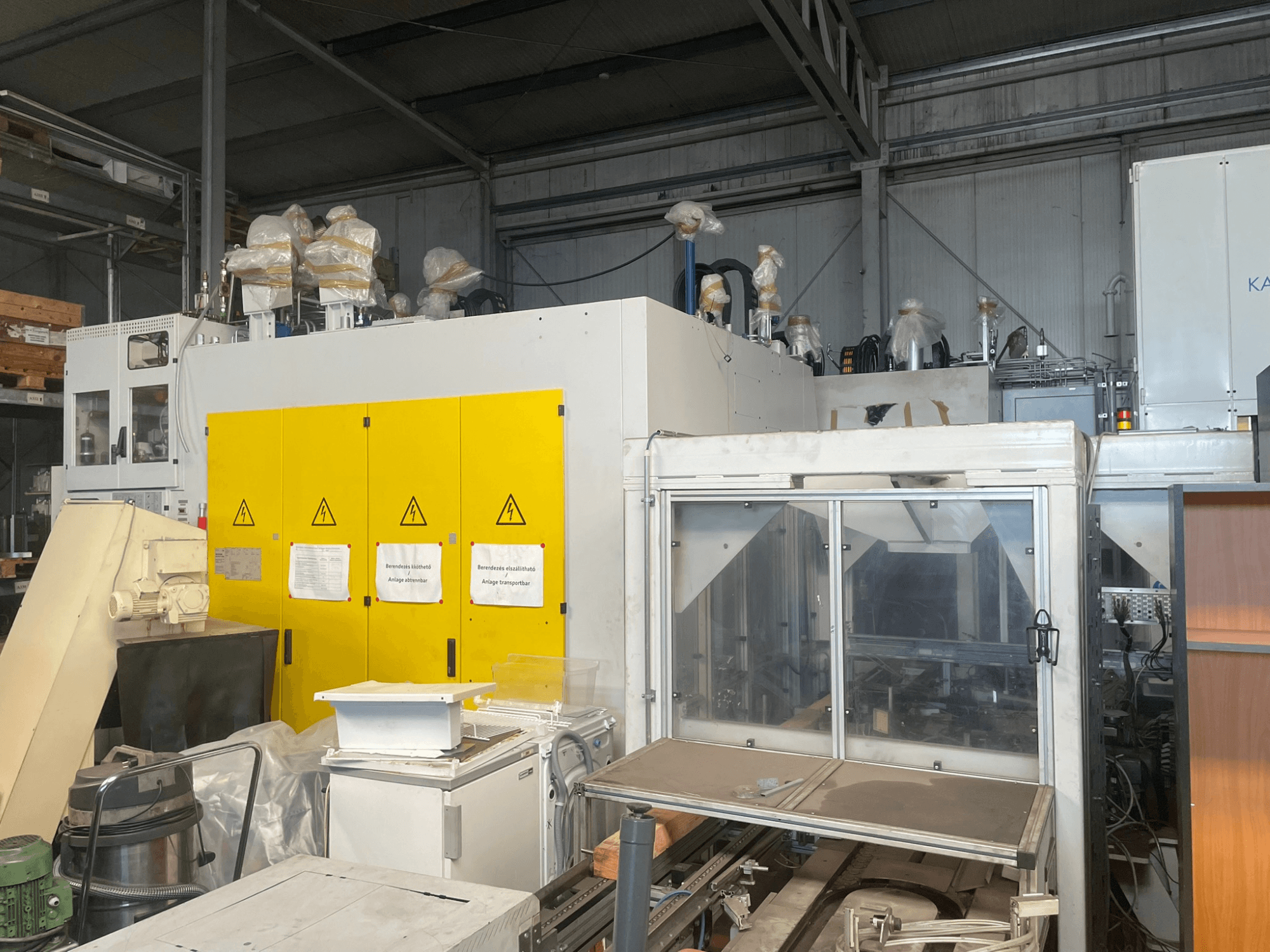 Maquinaria industrial con panel de control amarillo, vista lateral, con etiquetas de advertencia y cubiertas de plástico, en un entorno de almacén.