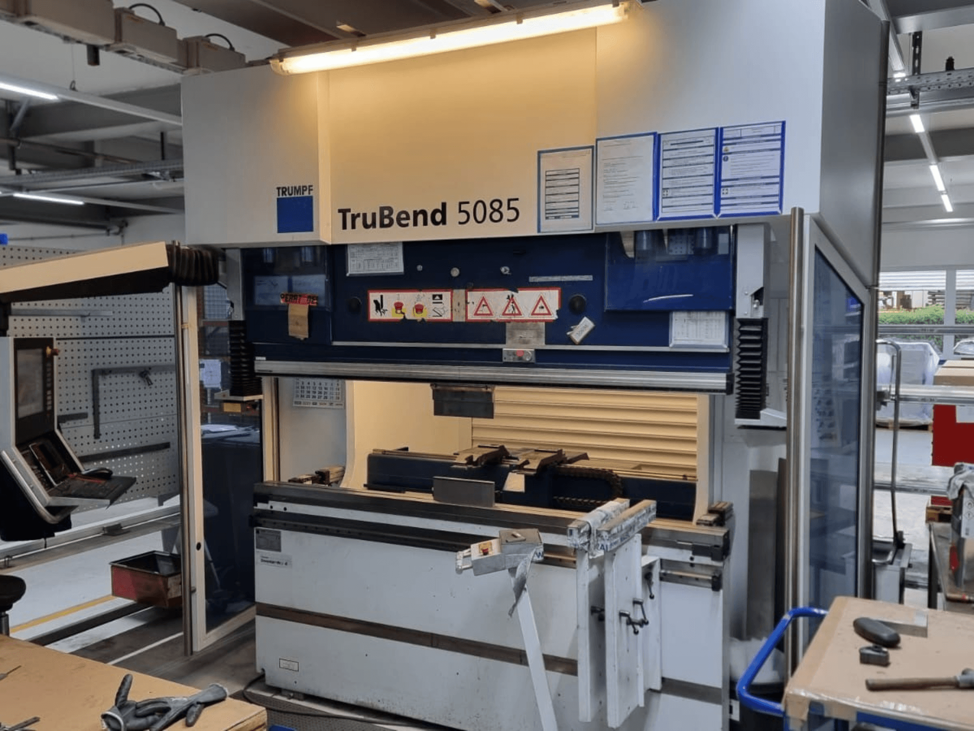Plegadora TRUMPF TruBend 5085, vista frontal, mostrando el panel de control, el puesto de mando y las advertencias de seguridad en el interior del taller.