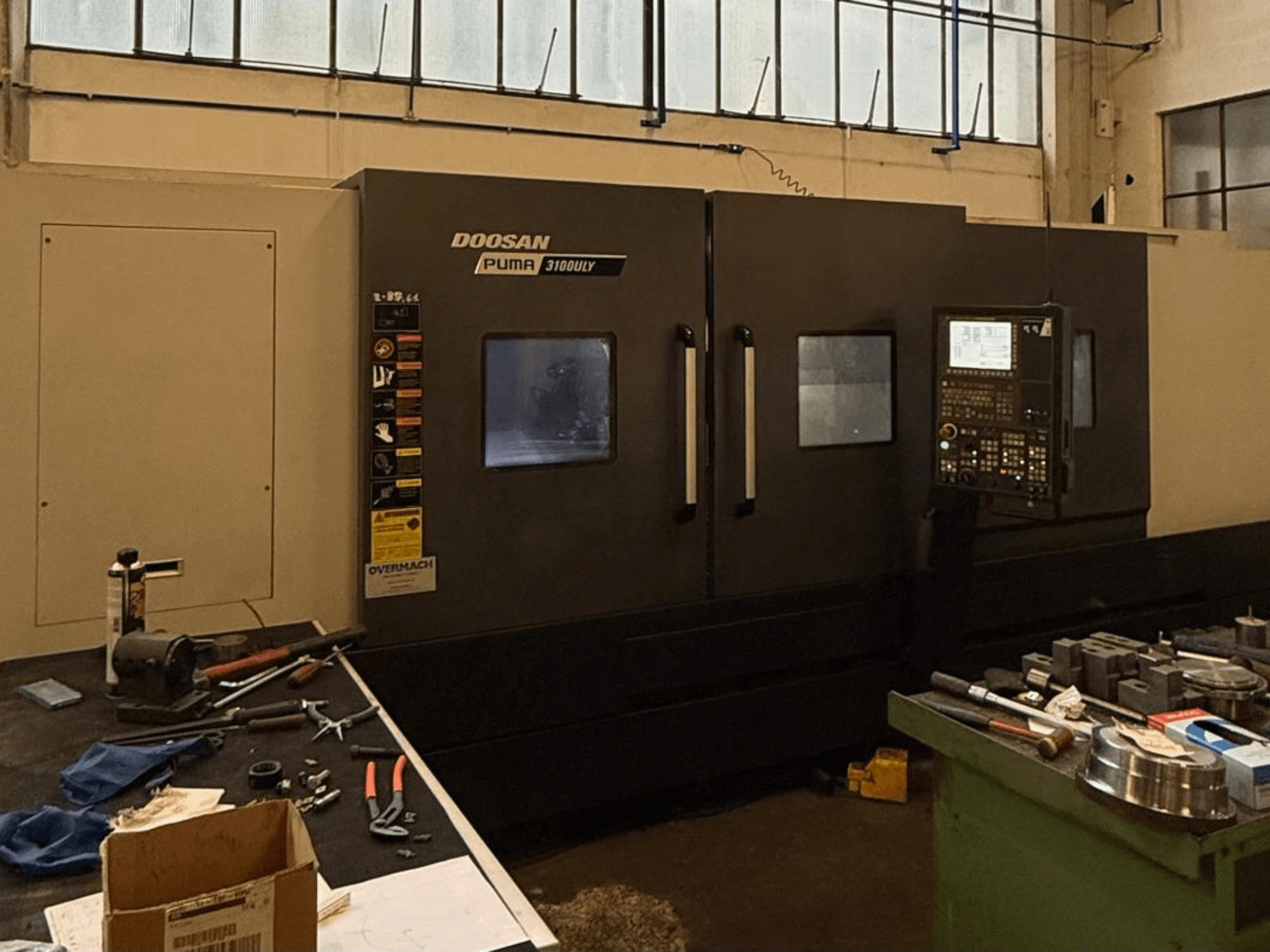 Torno CNC Doosan Puma 3100LY en un taller, vista lateral, con el panel de control y las herramientas visibles en el banco de trabajo.