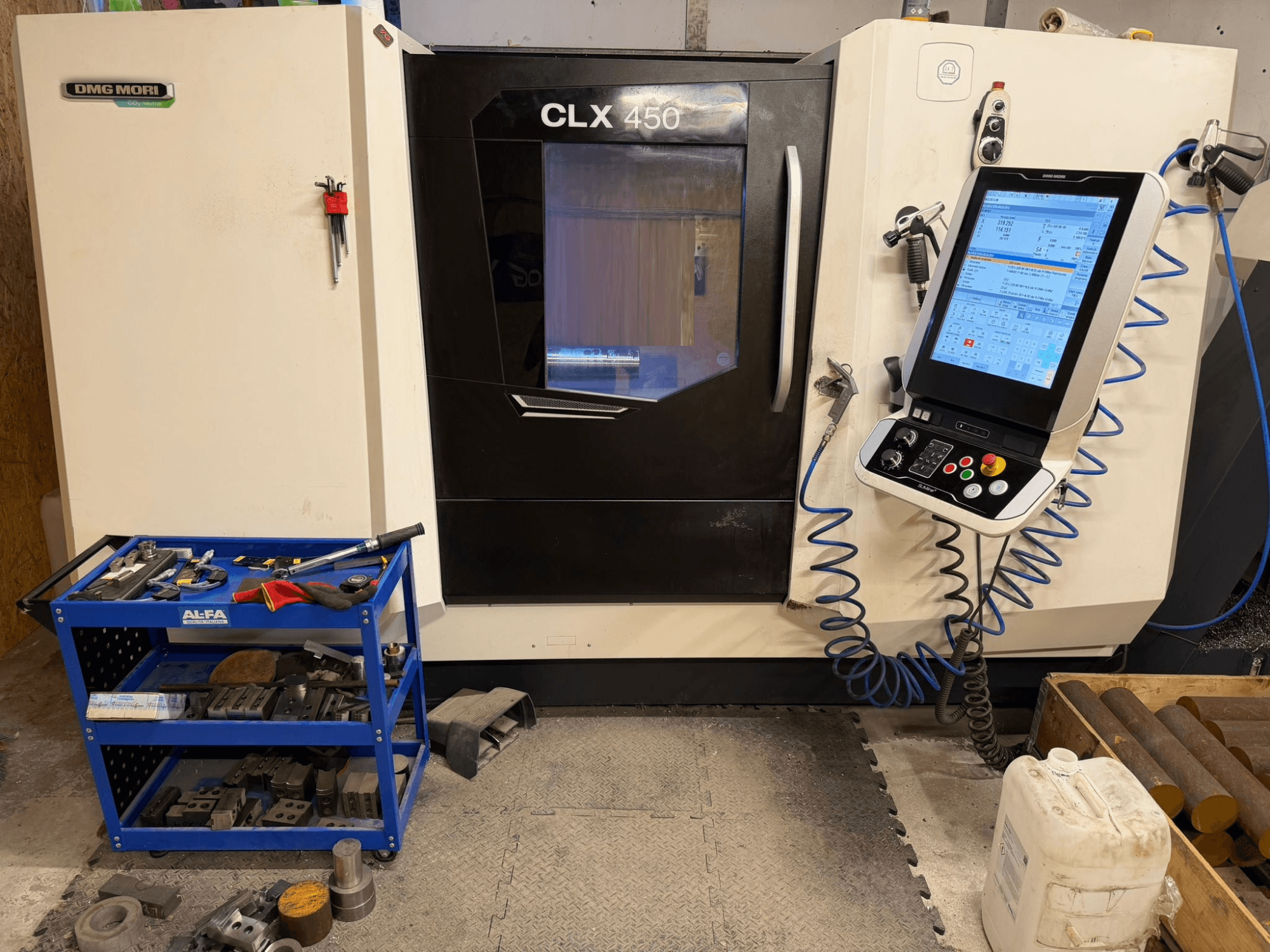 Vista frontal del torno horizontal DMG MORI CLX 450 V3, con un panel de control y un carro de herramientas cercano con equipamiento.