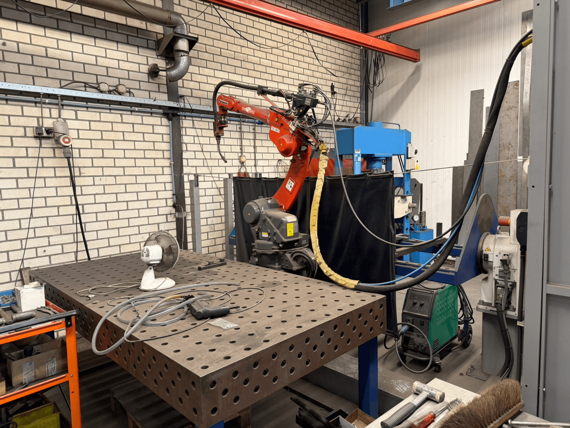 Vista frontal de un robot de soldadura PANASONIC TA-1800 en una estación de trabajo, con un ventilador y el equipo de soldadura visibles.