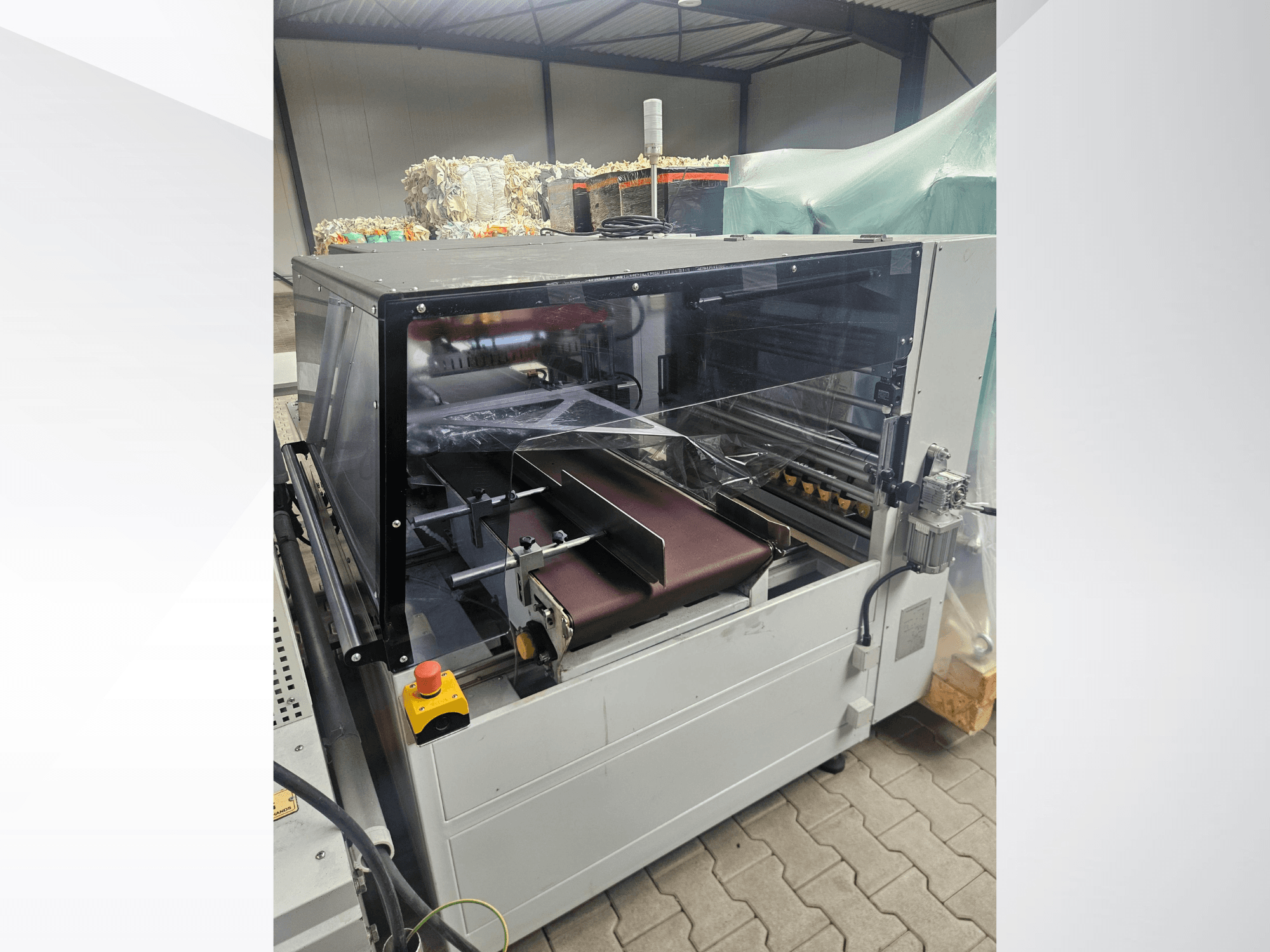 Máquina AMTEC Speed Shrink 40 PRO, vista frontal, en la que se aprecia un mecanismo de procesamiento de plástico con una cubierta de seguridad transparente y una cinta transportadora.