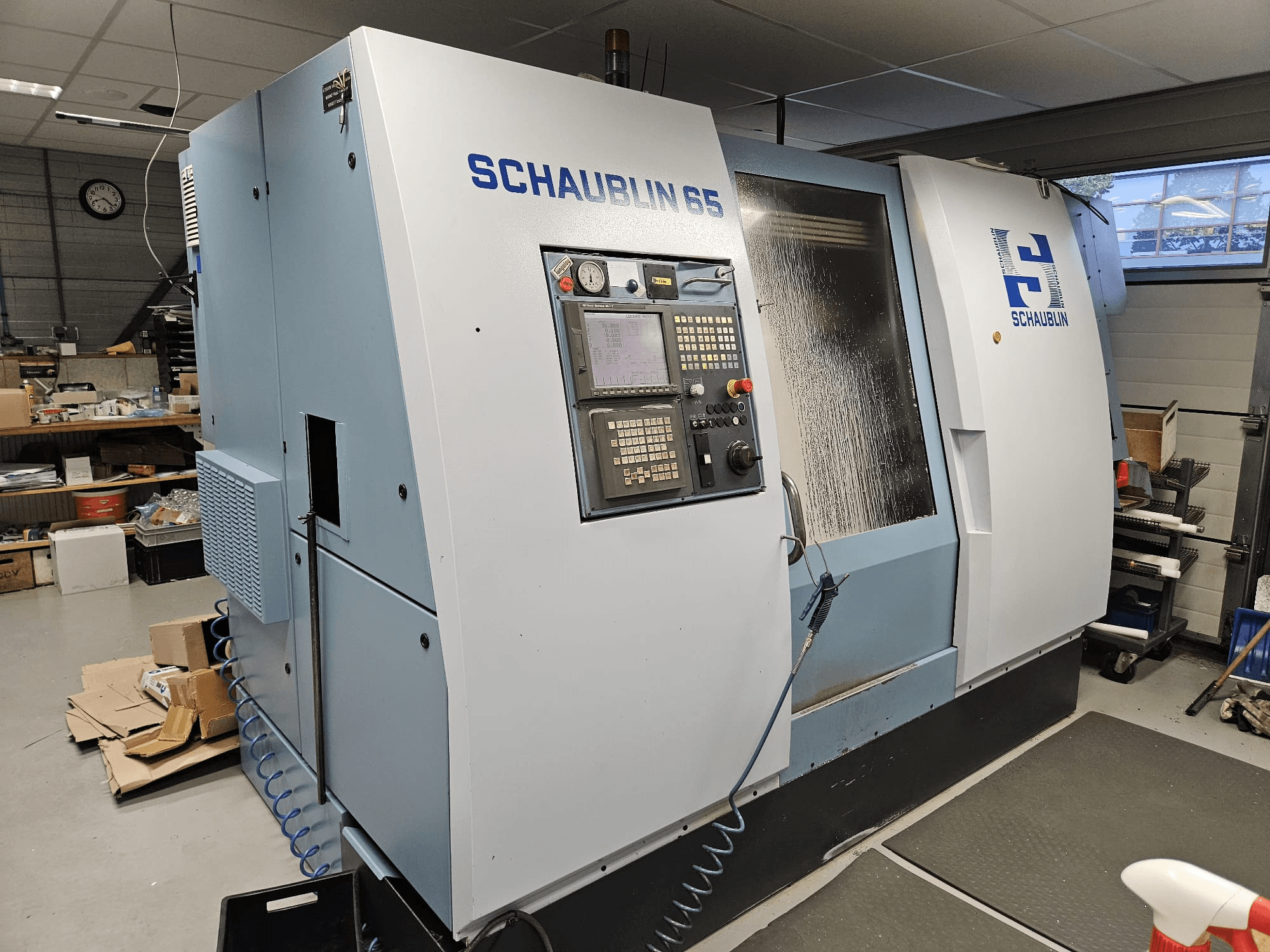 Máquina CNC SCHAUBLIN 65, vista lateral, con panel de control, carcasa azul y gris, y sistema de refrigerante visible.