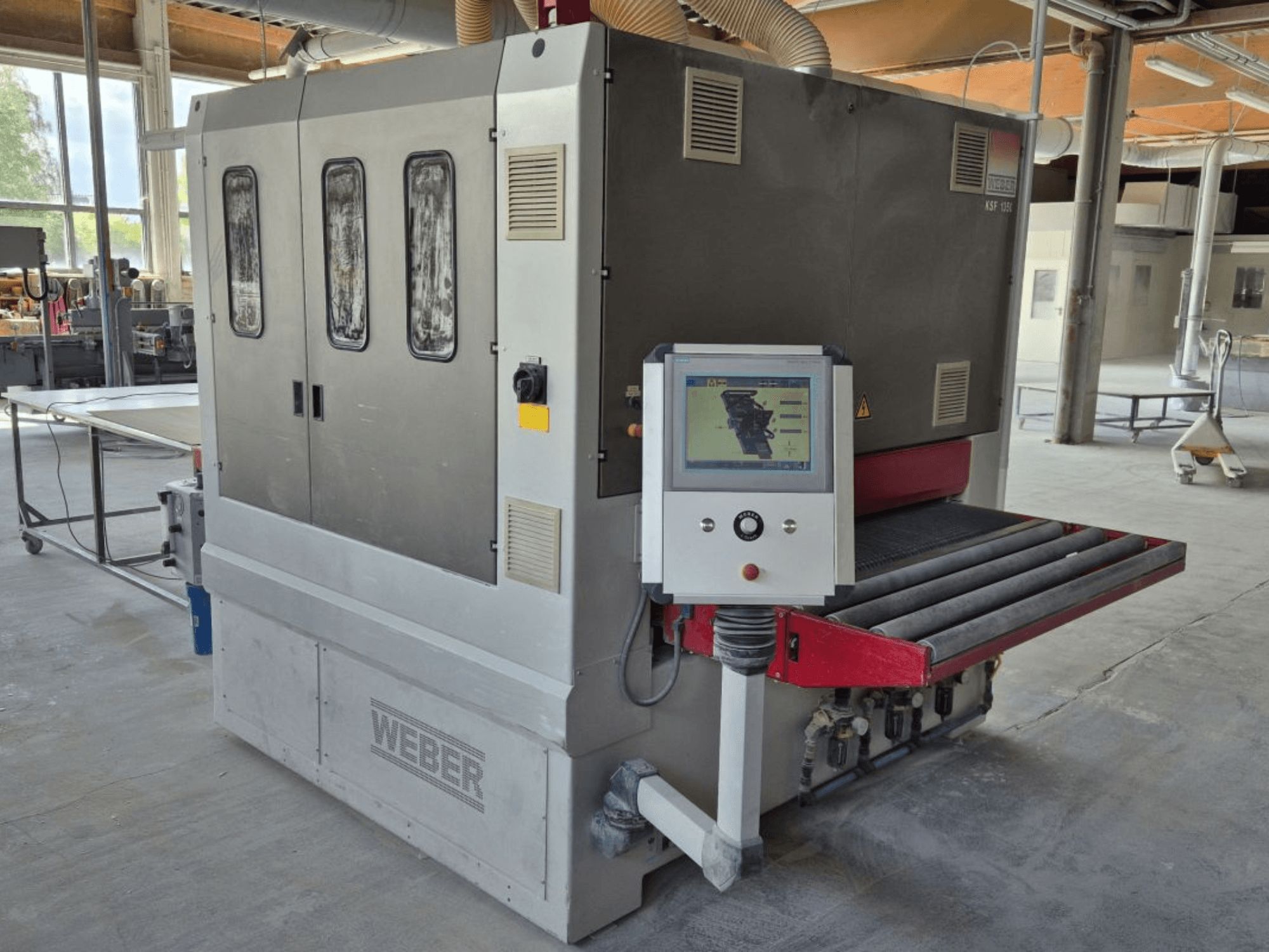 Lijadora de banda WEBER KSF-3-1350, vista lateral mostrando controles, panel de potencia y cinta de lijado. Entorno industrial con máquinas visibles.