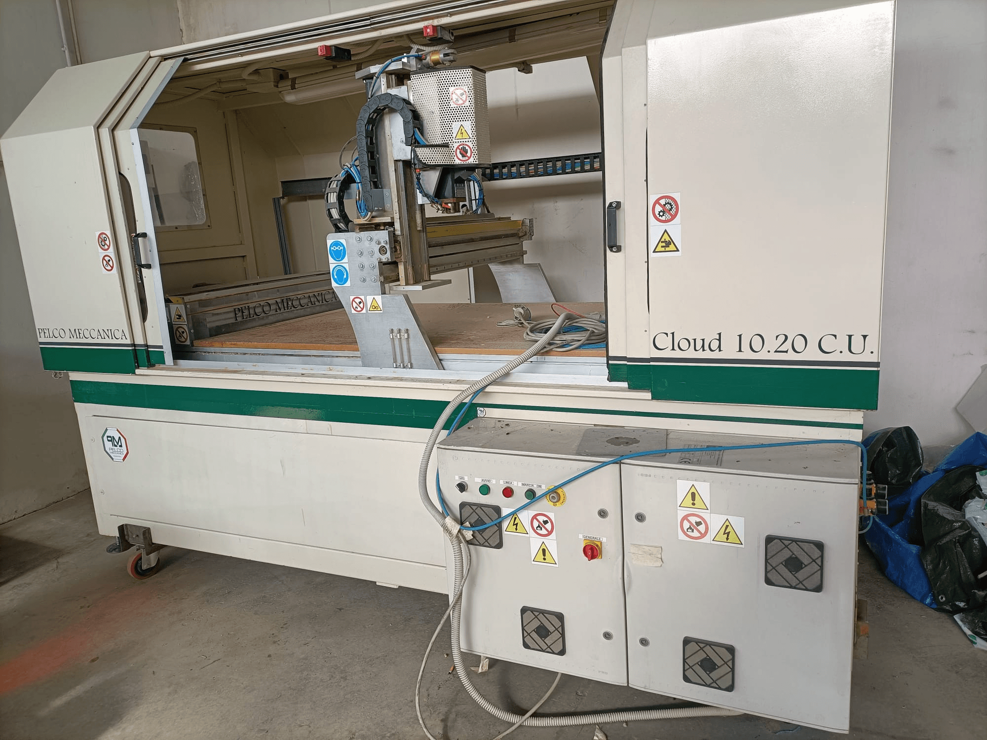 Ruteadora CNC MECCANICA CLOUD 10.20 C.U., vista frontal mostrando el panel de control y el espacio de trabajo, con etiquetas de seguridad y mangueras visibles.