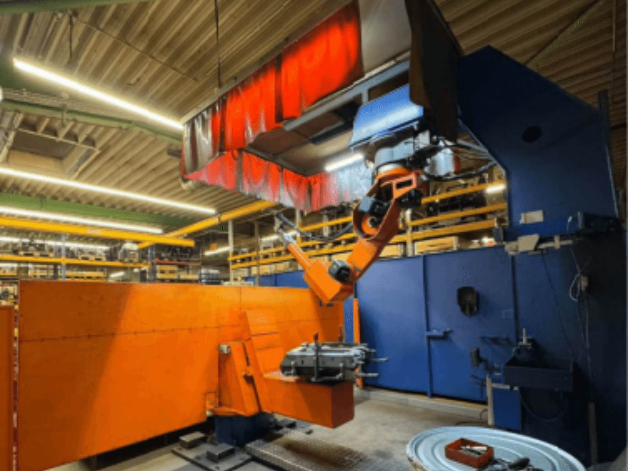 Robot de soldadura CLOOS Romat 350 e en un entorno industrial, vista frontal con brazo naranja, sujetando una pieza de trabajo bajo una cortina superior.