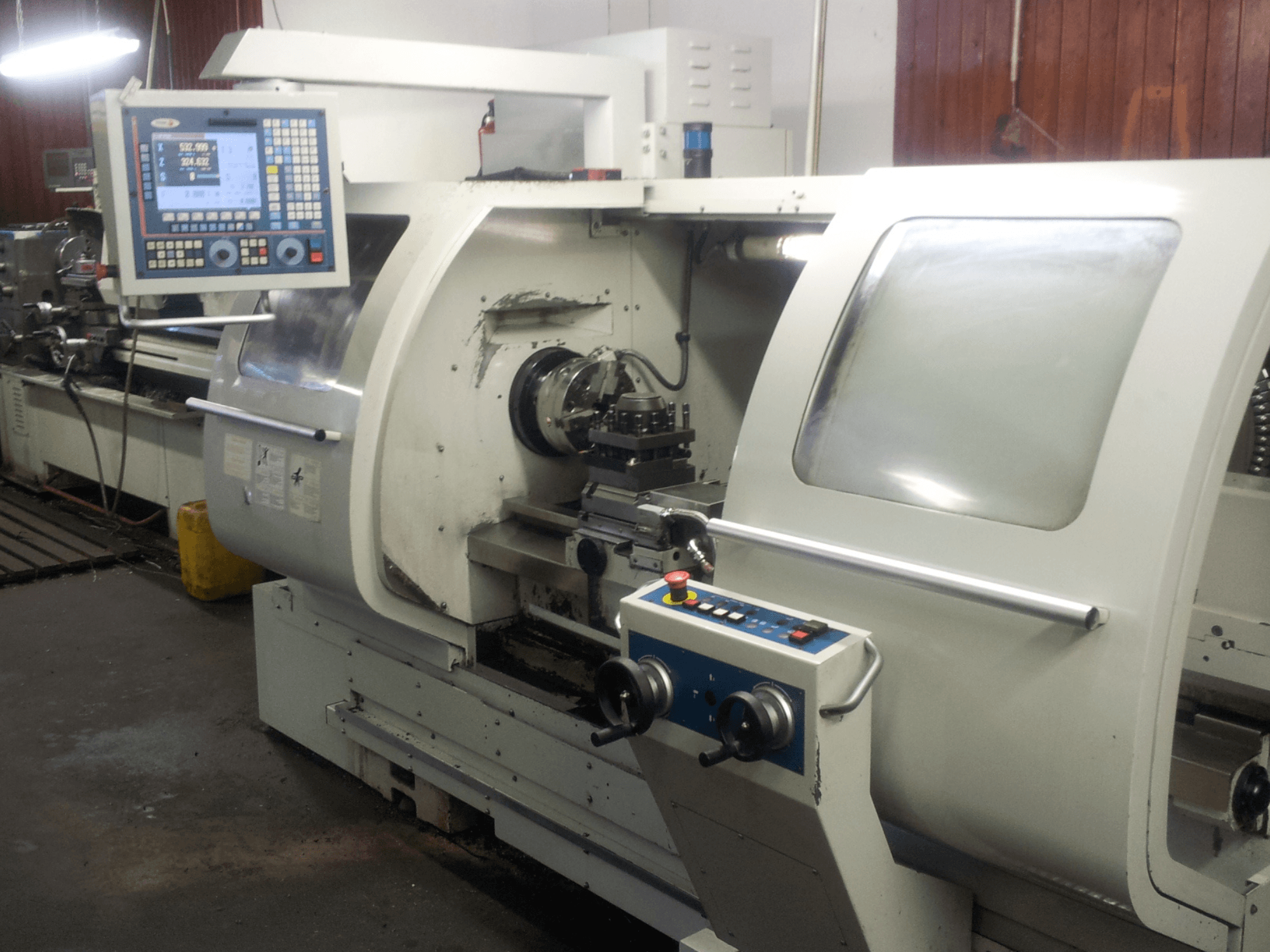 Vista frontal de un torno CNC con panel de control digital, destacando el plato, el husillo y los dispositivos de seguridad.