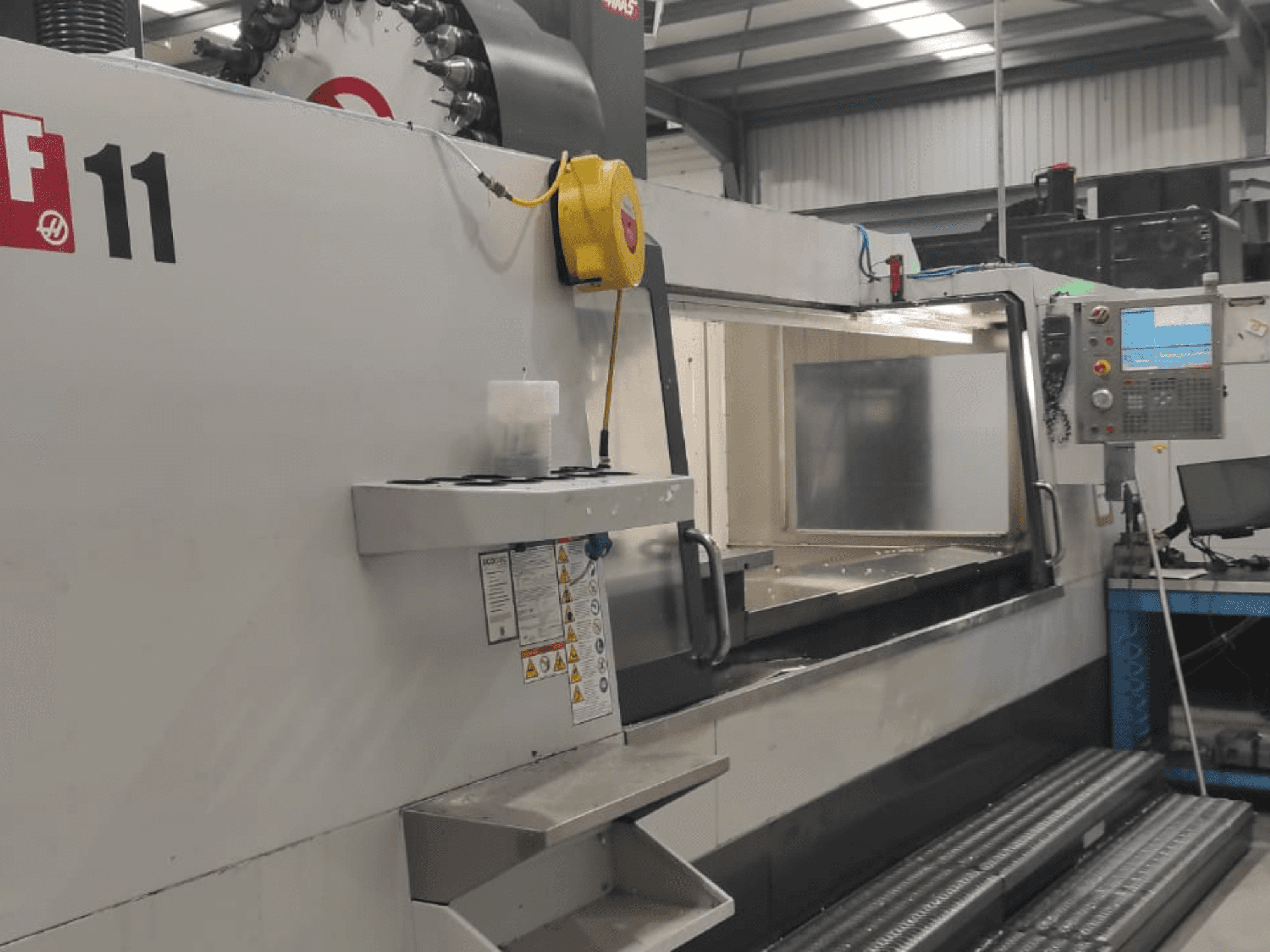 Maquinaria de Haas, modelo VF-11, mostrada desde una vista frontal con panel de control, amplia zona de trabajo y etiquetas de seguridad visibles.