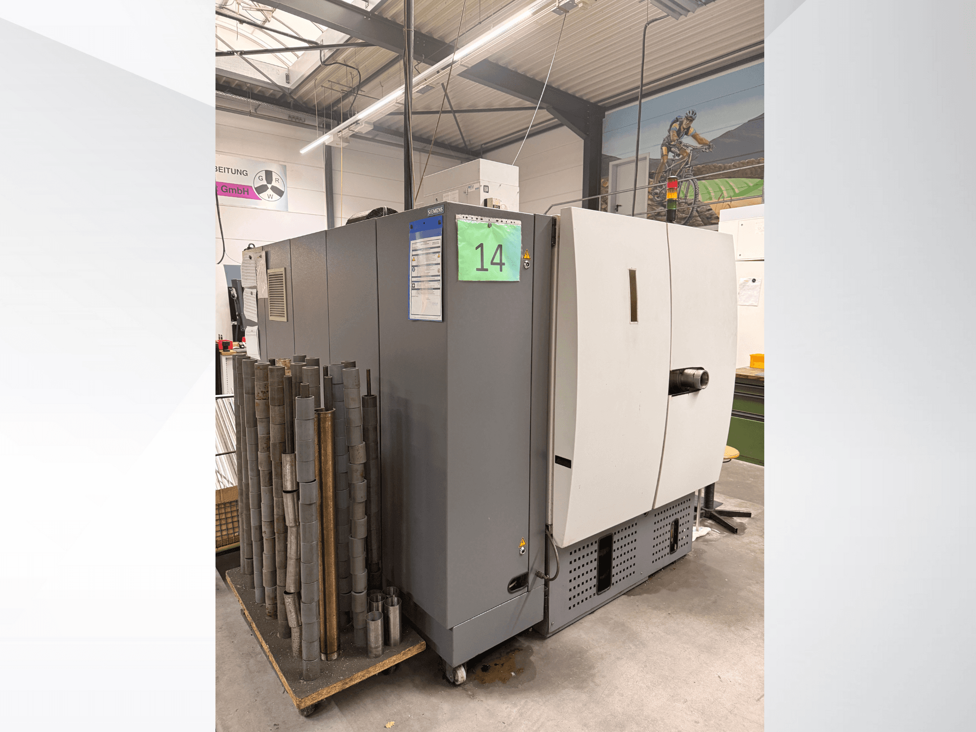 Máquina industrial gris de Siemens, vista lateral, con existencias de piezas metálicas cilíndricas a su lado y etiquetas de equipo visibles.