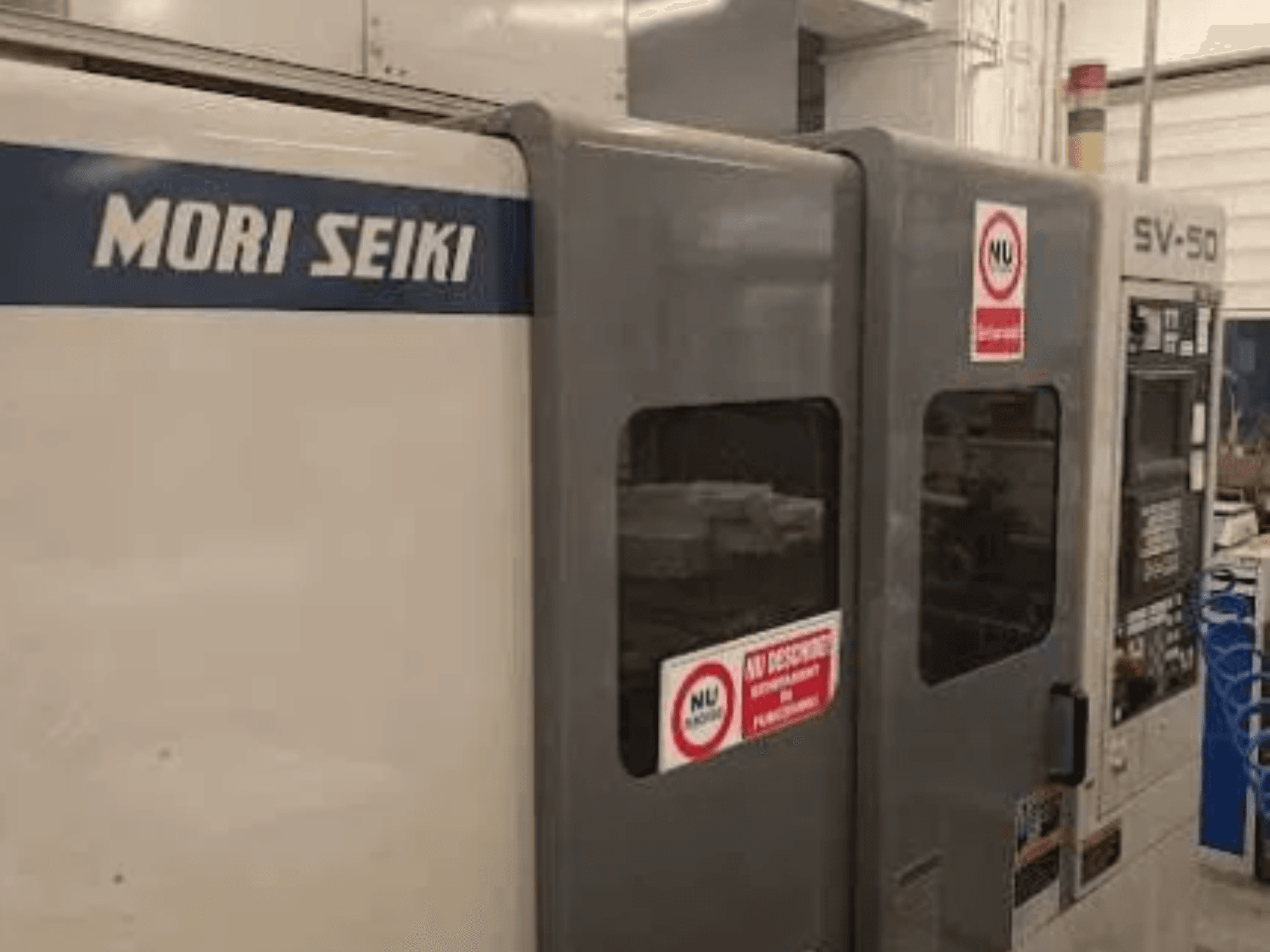 Máquina CNC MORI SEIKI SV-50, vista lateral; presenta panel de control y adhesivos de advertencia de seguridad.
