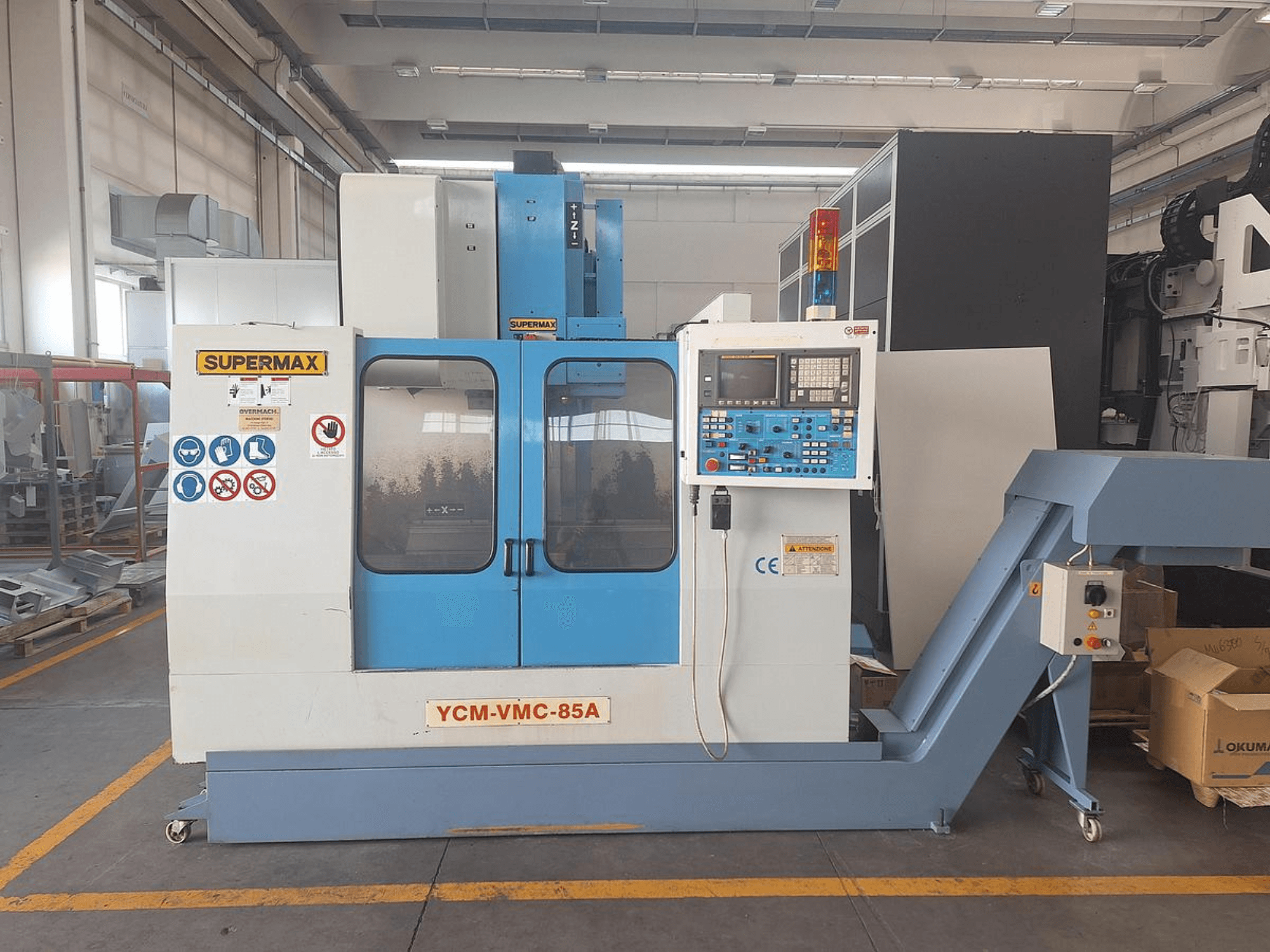 Centro de mecanizado vertical SUPERMAX YCM-VMC-85A en una fábrica, con panel de control y adhesivos de seguridad; vista frontal.