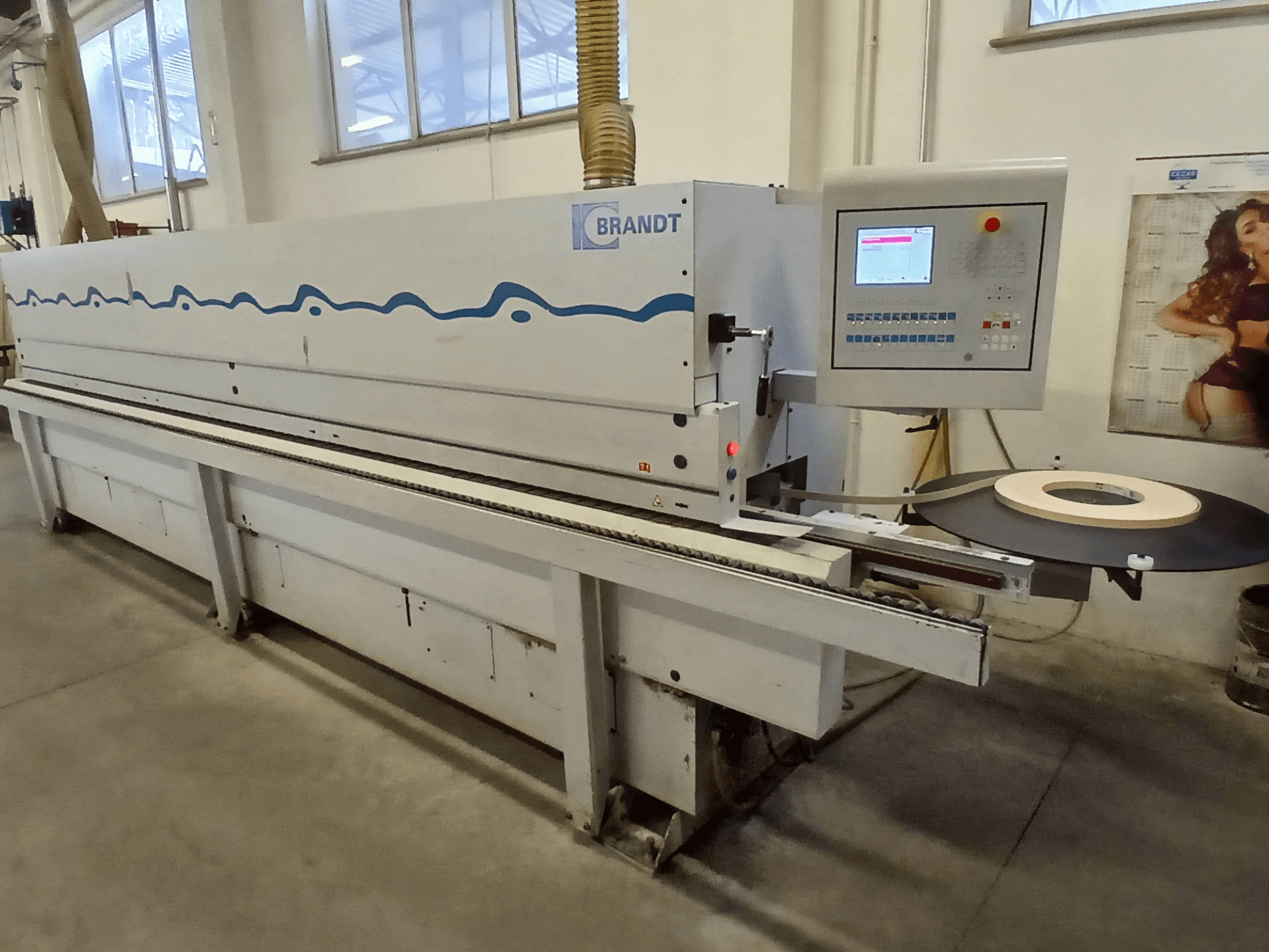 Encoladora de cantos Brandt, vista lateral, con un panel de control y un rollo de cinta en la mesa de alimentación.