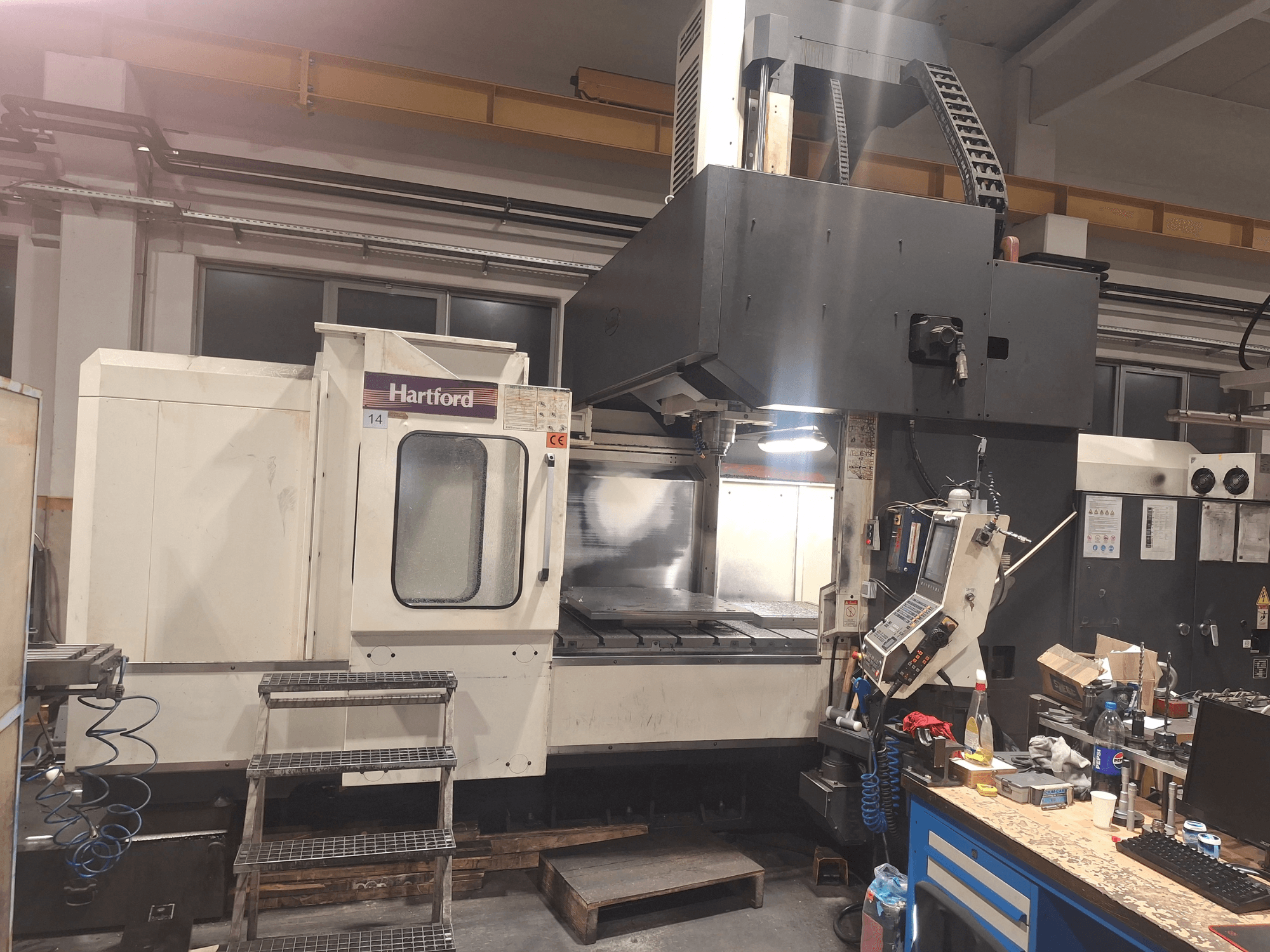 Máquina CNC Hartford, vista frontal, con controles técnicos, escalones de acceso y zona de mesa de trabajo, en un entorno de taller.