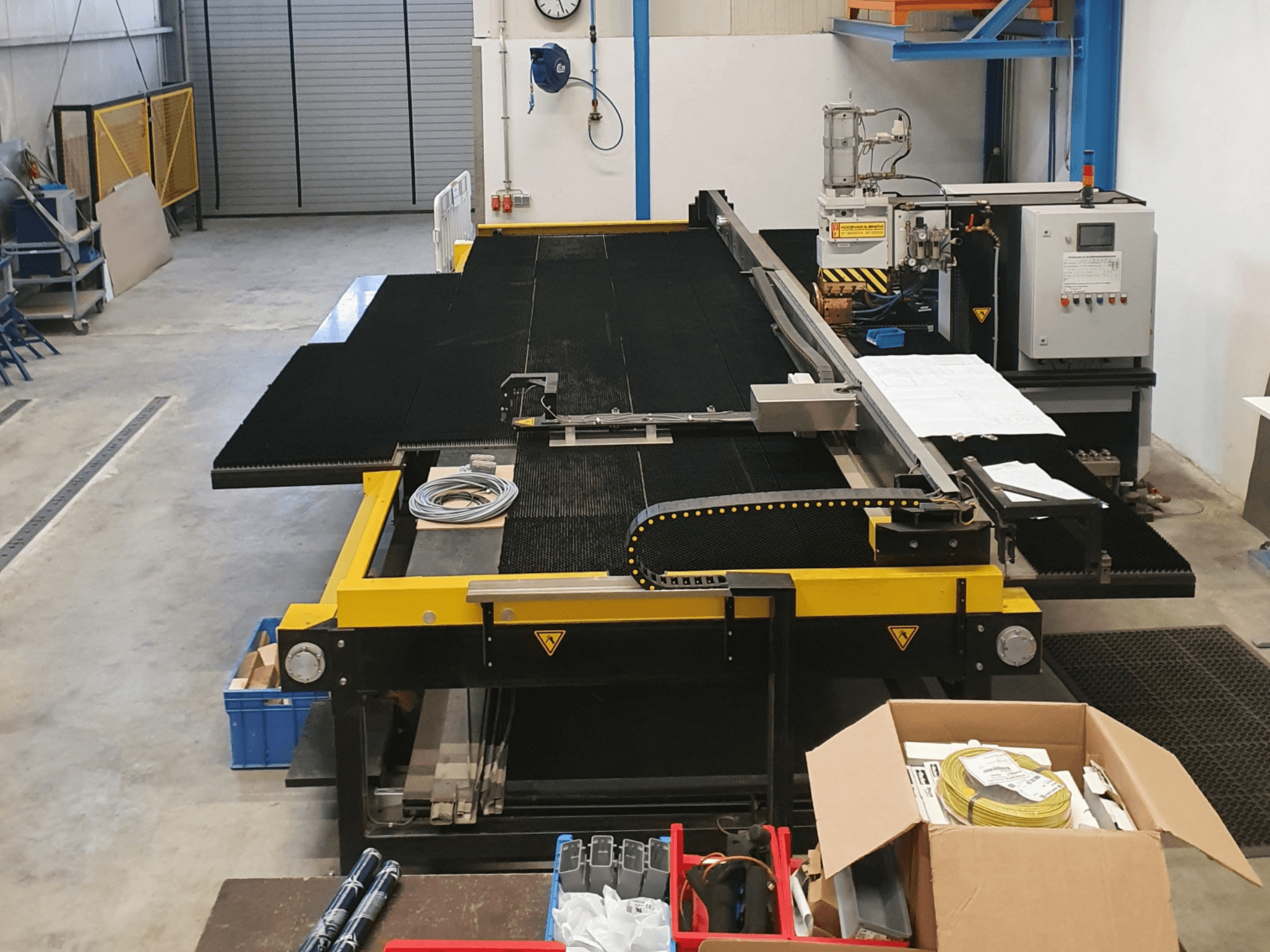 Máquina de corte industrial con características amarillas y negras, vista frontal, mostrando controles y accesorios en un entorno de fábrica.