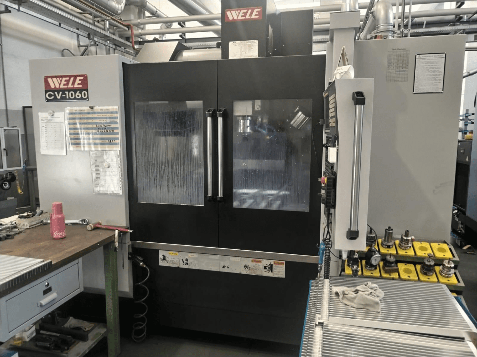 Centro de mecanizado vertical Wele CV-1060, vista frontal, con panel de control y área de trabajo visible; incluye herramientas accesorias y etiquetas de seguridad.